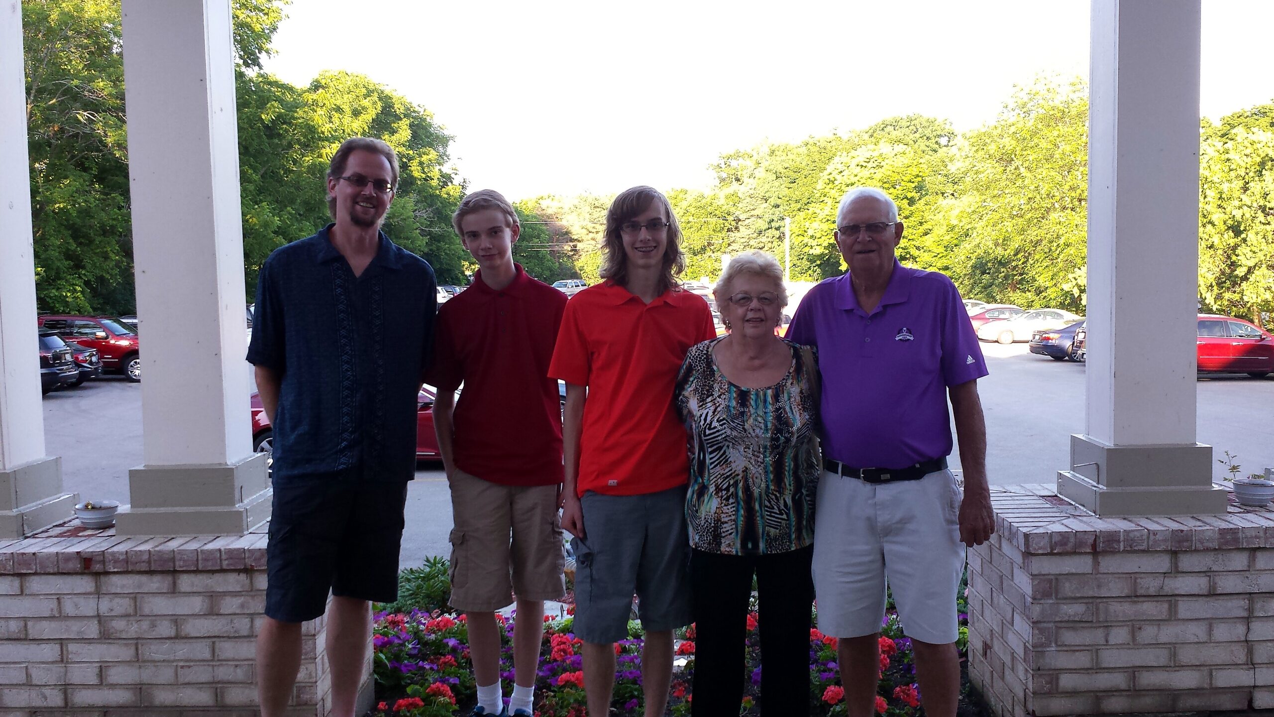 Five people posing together outdoors on a porch, smiling at the camera, with a flower bed in front and cars in the background.