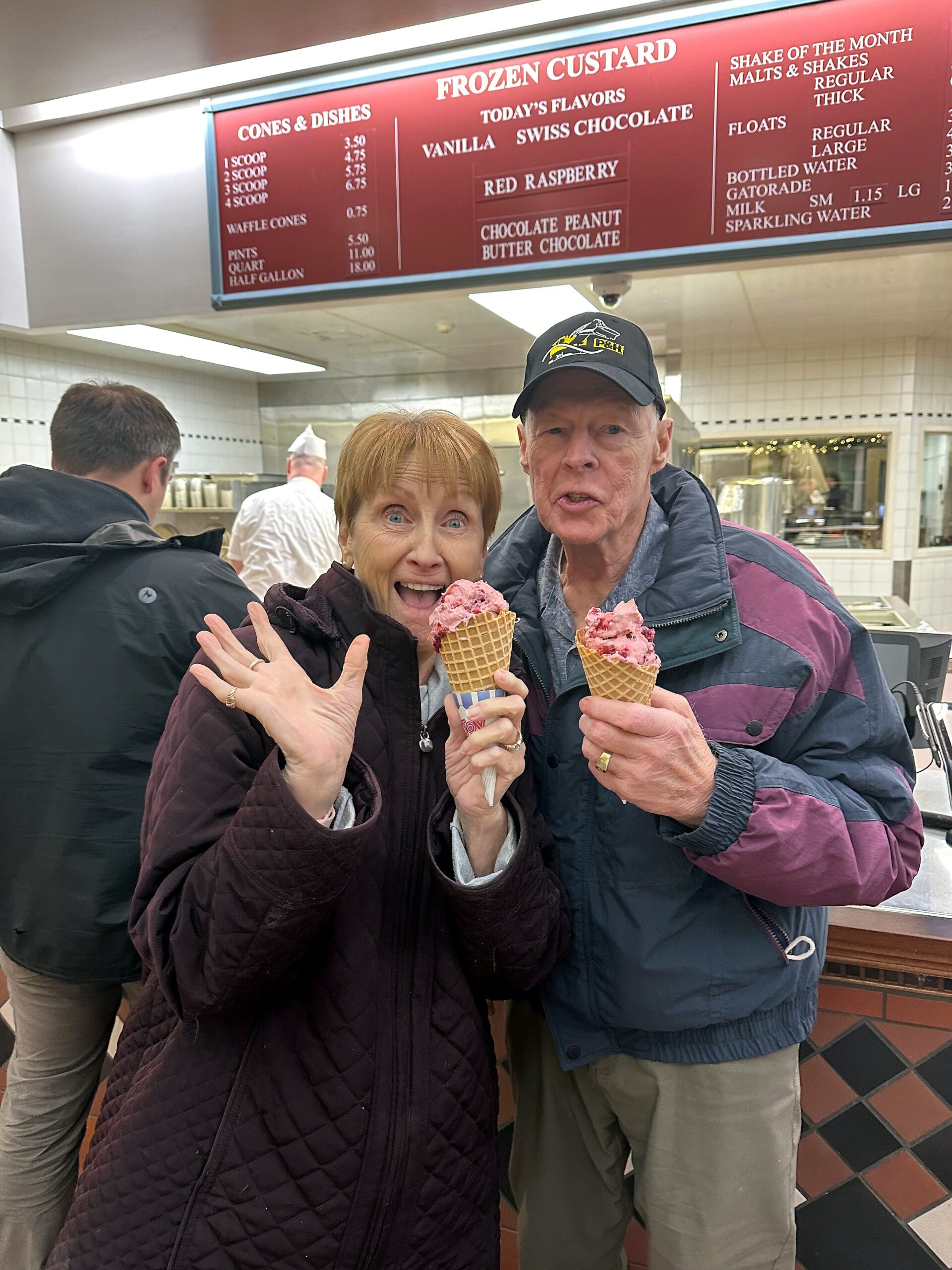 Elderly couple smiling and holding pink ice cream cones inside a frozen custard shop. Behind them is a red menu board and kitchen area.