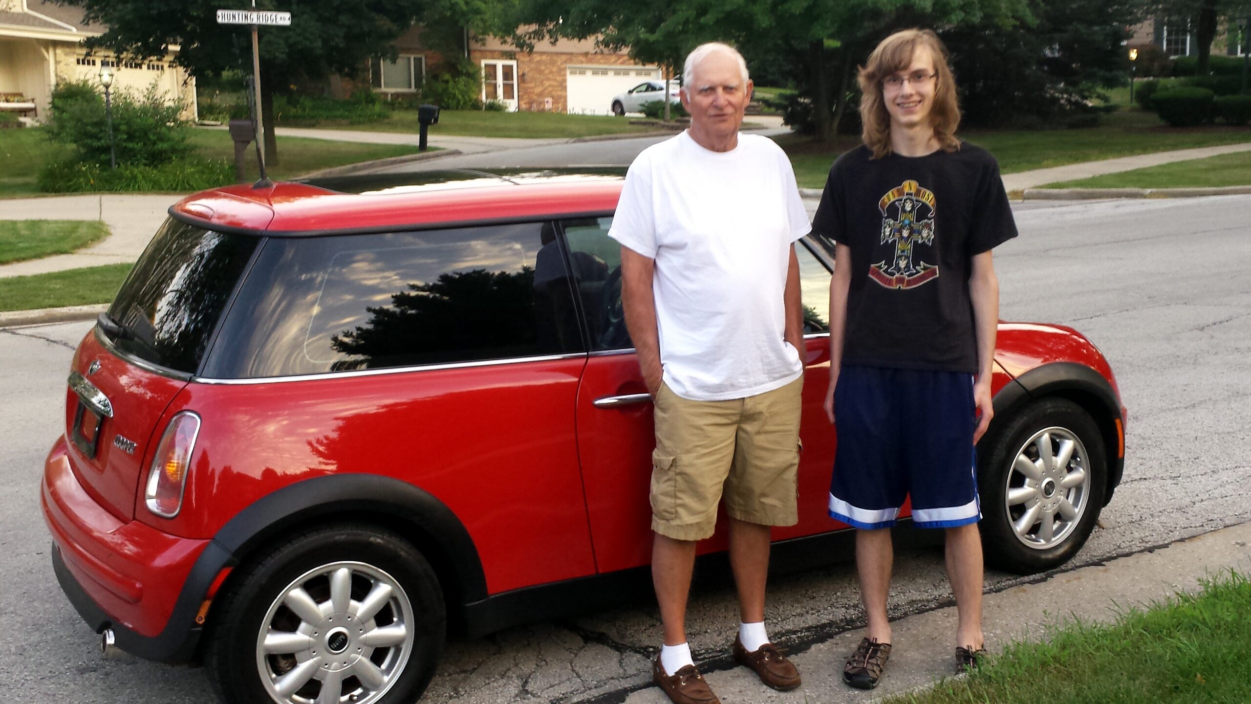 Two men pose beside a red car parked on a suburban street; an older man in a white t-shirt and khaki shorts stands next to a younger man with long hair and a black graphic t-shirt and blue shorts.