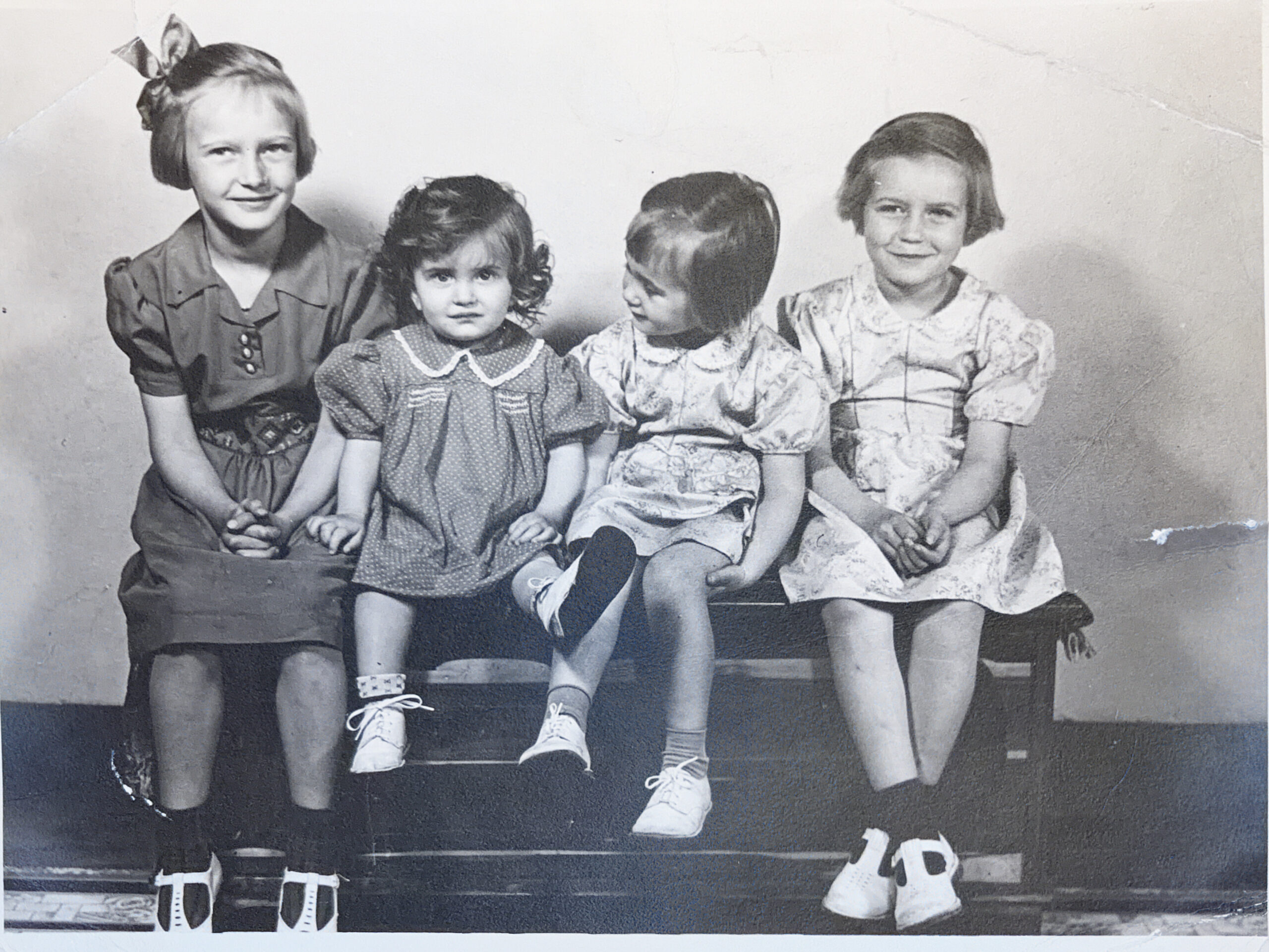 Four young girls in vintage dresses sit on a bench for a photo; the girl on the far left has a bow in her hair.