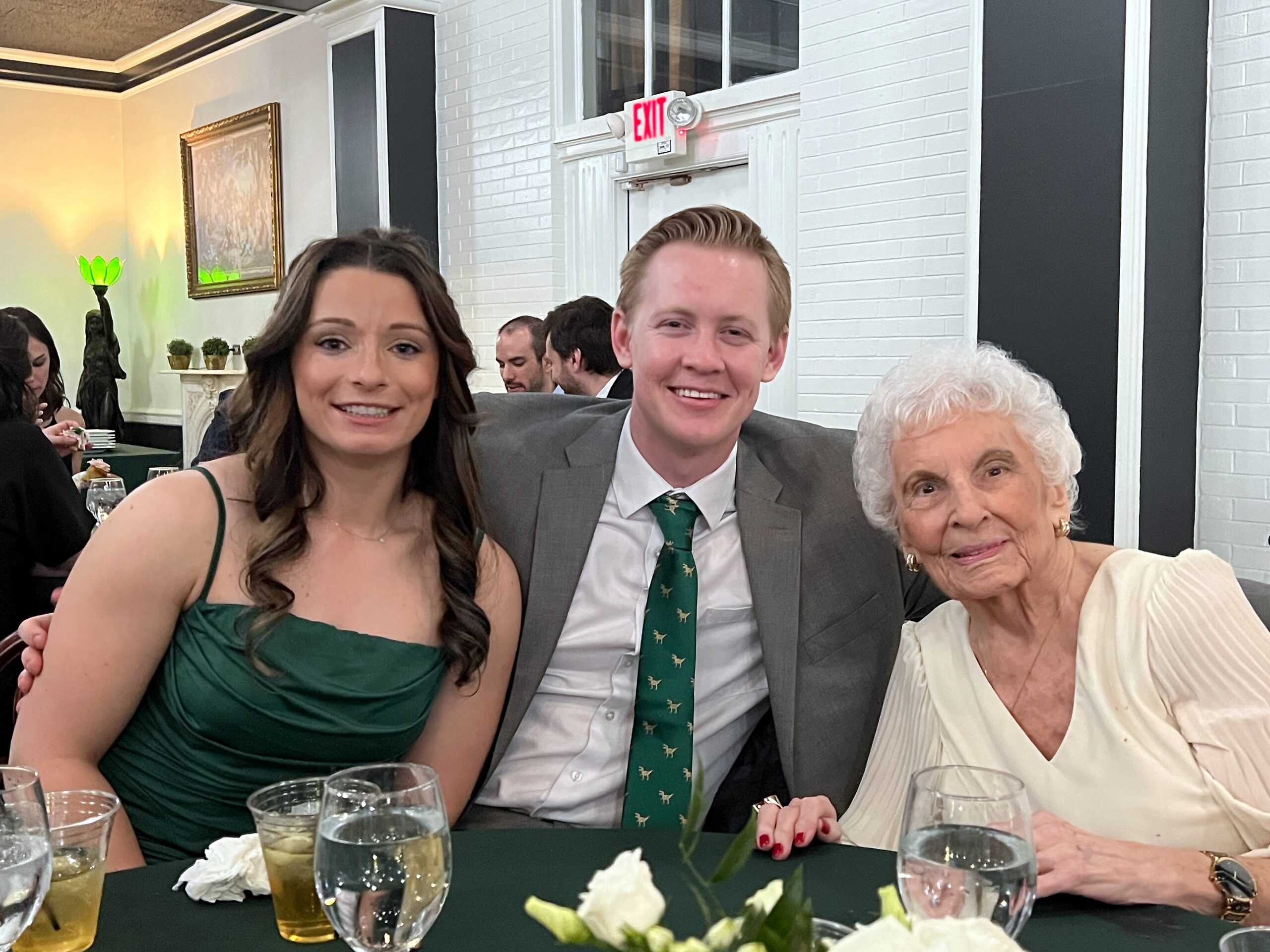 Group of three guests in formal wear posing at a dining table with drinks and a floral centerpiece.