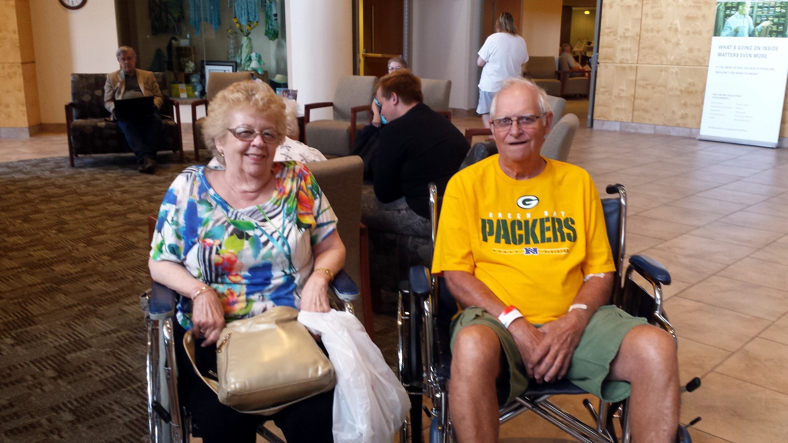 Smiling elderly couple in wheelchairs sit side by side in a hospital lobby, woman in a colorful blouse and man in a Packers shirt, with friends and reception area behind them.