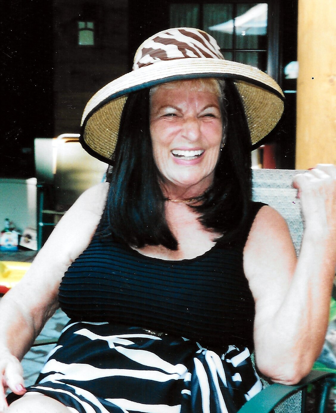 Smiling elderly woman wearing a wide-brimmed striped hat and a black striped dress, seated outdoors.