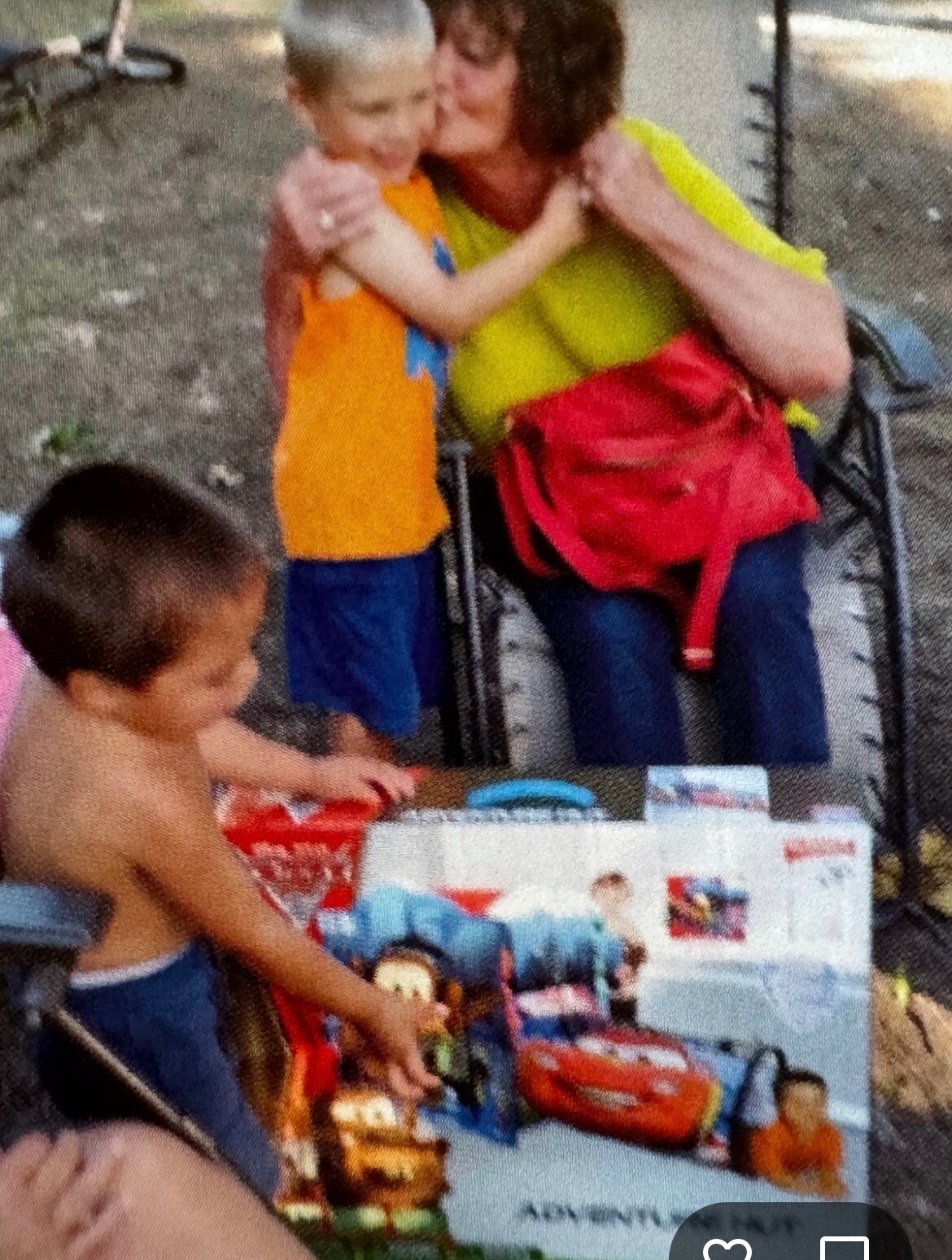 A woman in a yellow top hugs a child in an orange shirt outdoors near a table of Toys and books for kids.