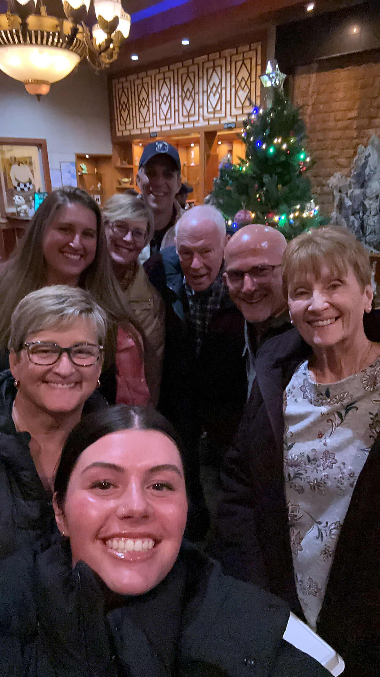 Group of seven people smiling for a selfie in a cozy restaurant with a decorated Christmas tree in the background.
