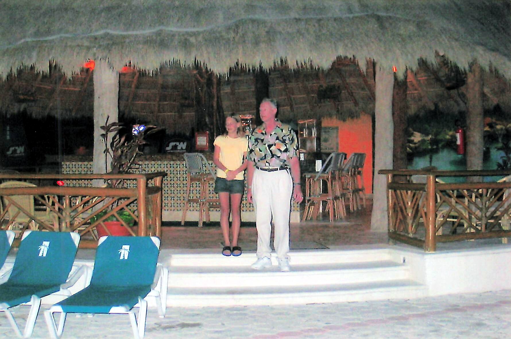 Two people, a man in a patterned shirt and white pants and a girl in a yellow shirt and shorts, pose on a pool deck at a tropical resort.