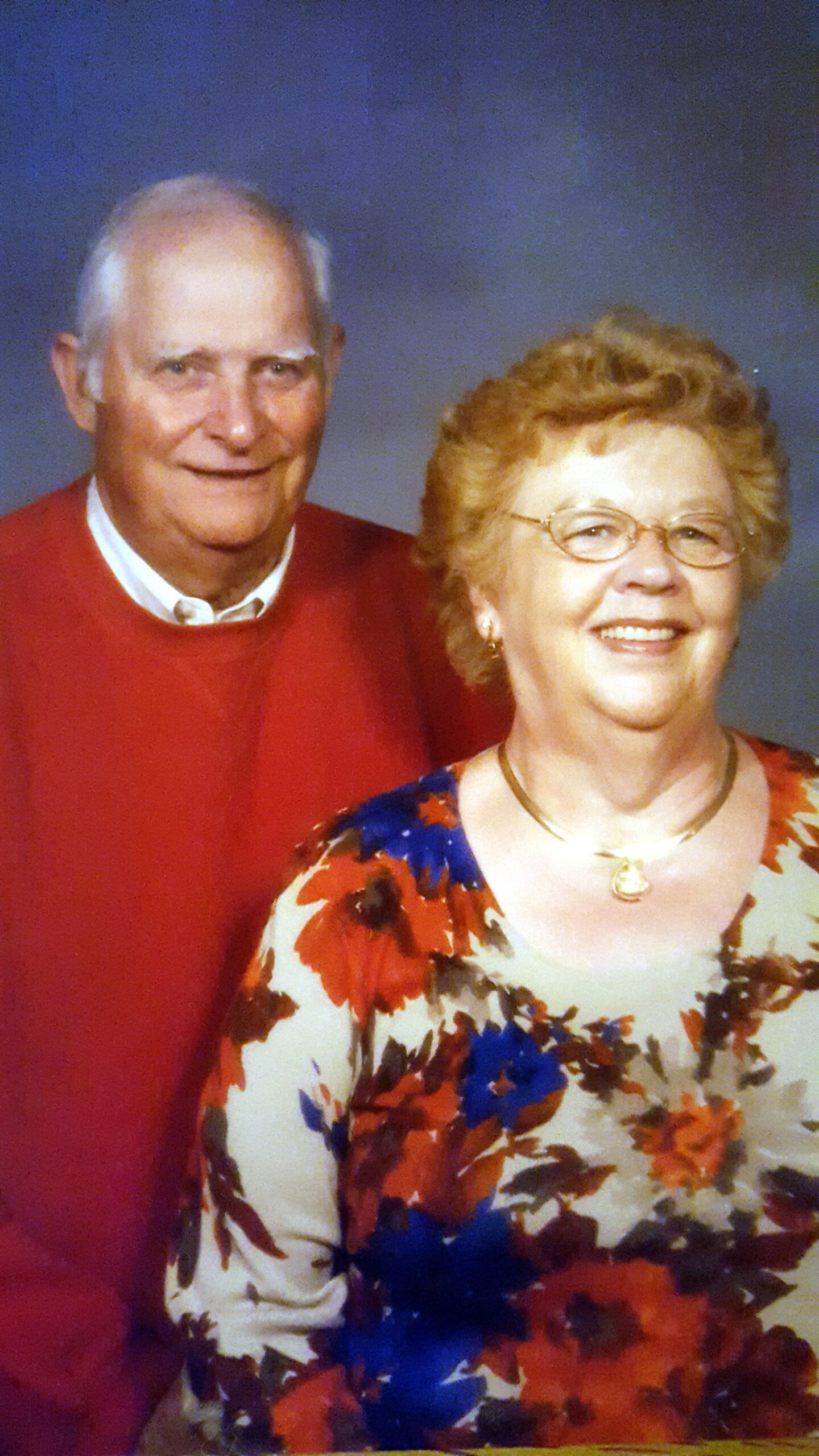 Smiling elderly couple posing together, man in red sweater and white collar, woman in floral top with glasses and necklace.