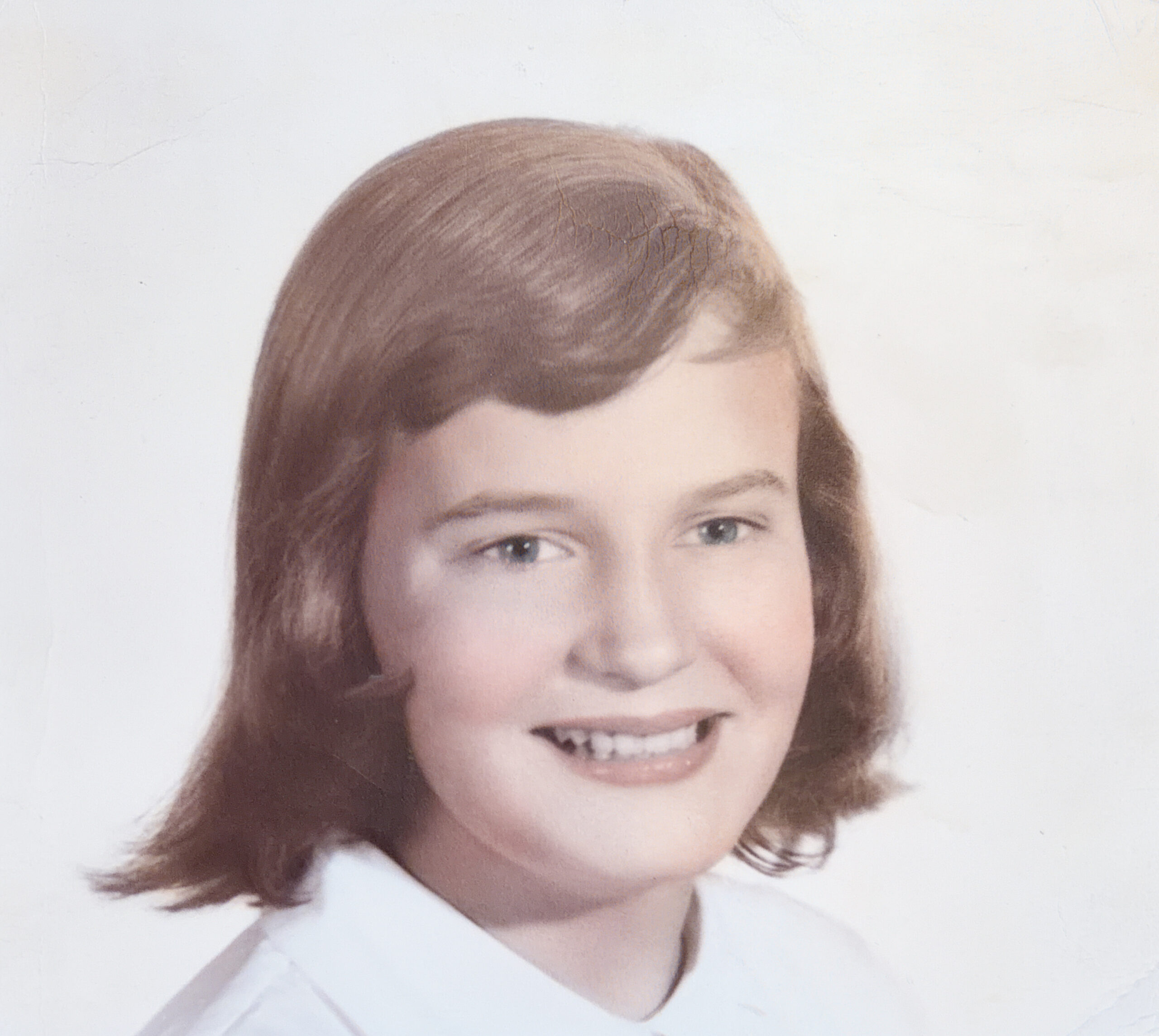 Close-up portrait of a smiling young girl with brown hair against a light background, facing the camera.