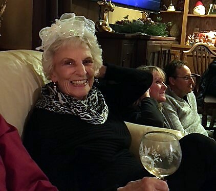 Smiling elderly woman wearing a white hat and leopard scarf sits on a sofa, holding a wine glass, with a festive living room and other guests in the background.
