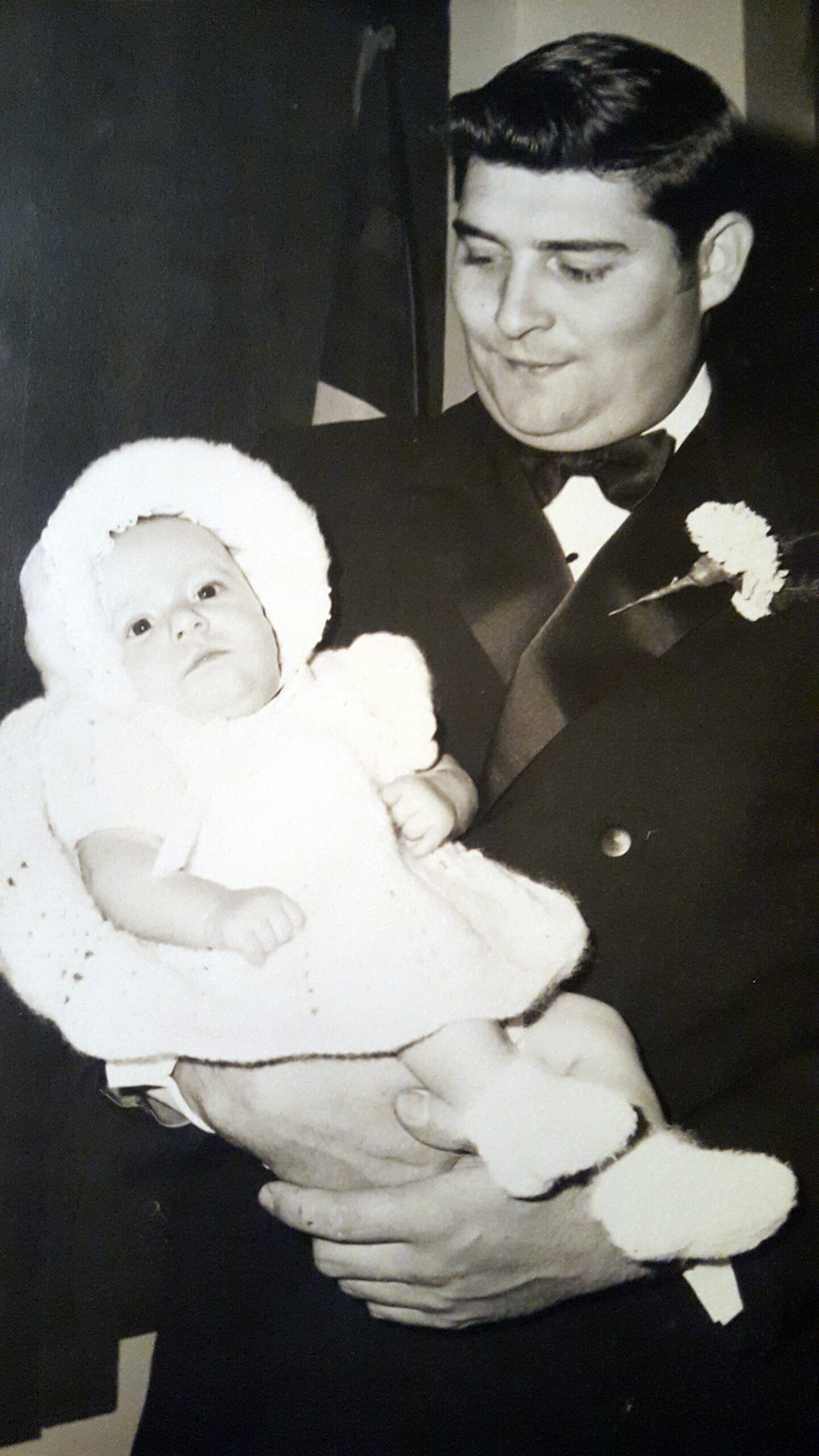 Man in a formal suit holding a small baby dressed in white, zippy bonnet, both looking gentle to one side.
