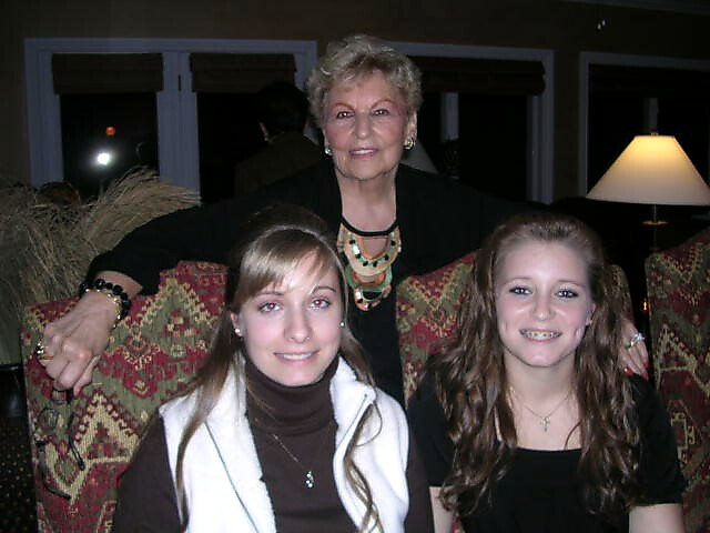 Older woman standing behind two young women seated on a patterned sofa in a living room, smiling at the camera.
