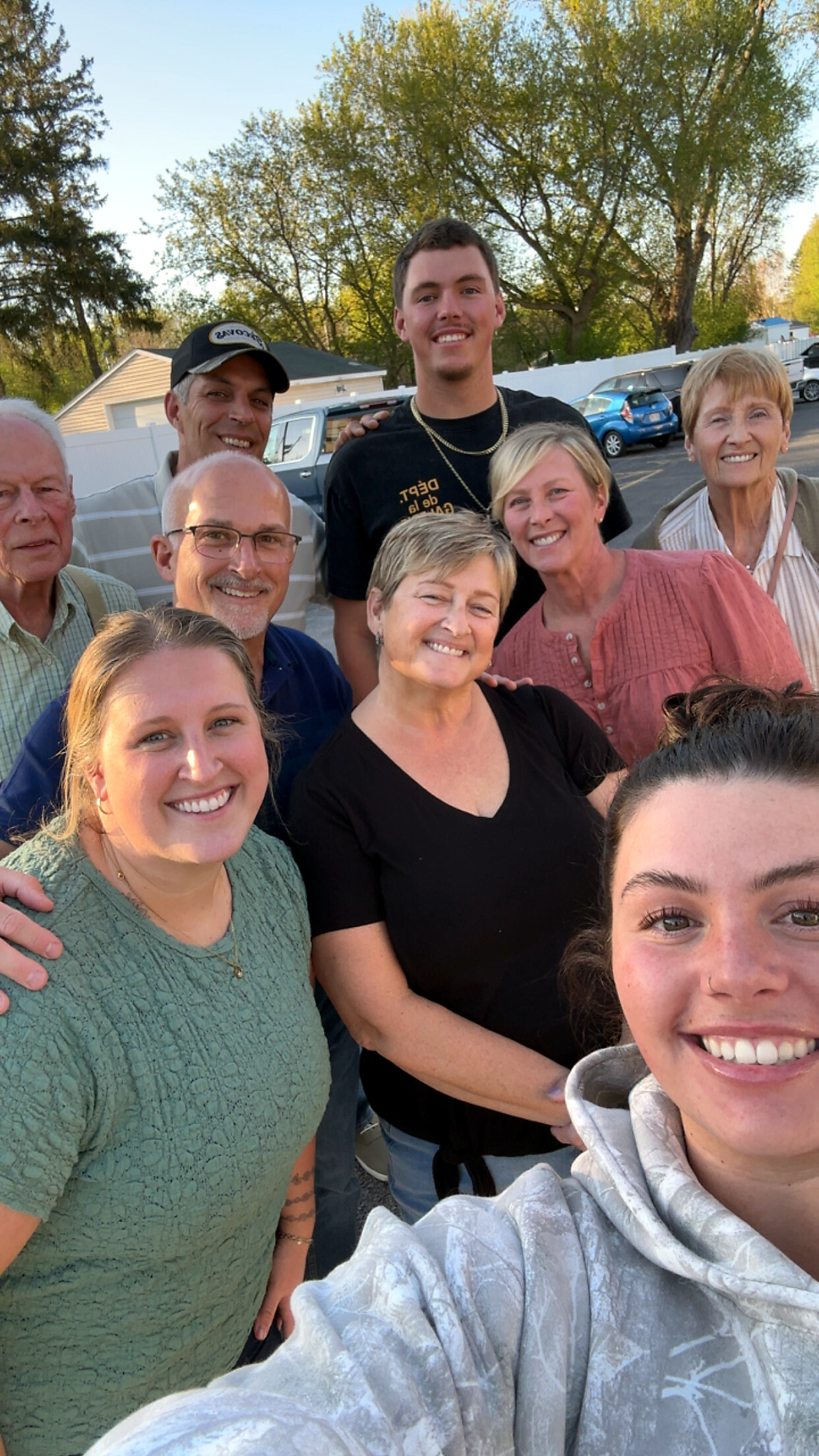 Group of eight adults posing for a selfie outdoors in a parking lot with trees in the background, all smiling.