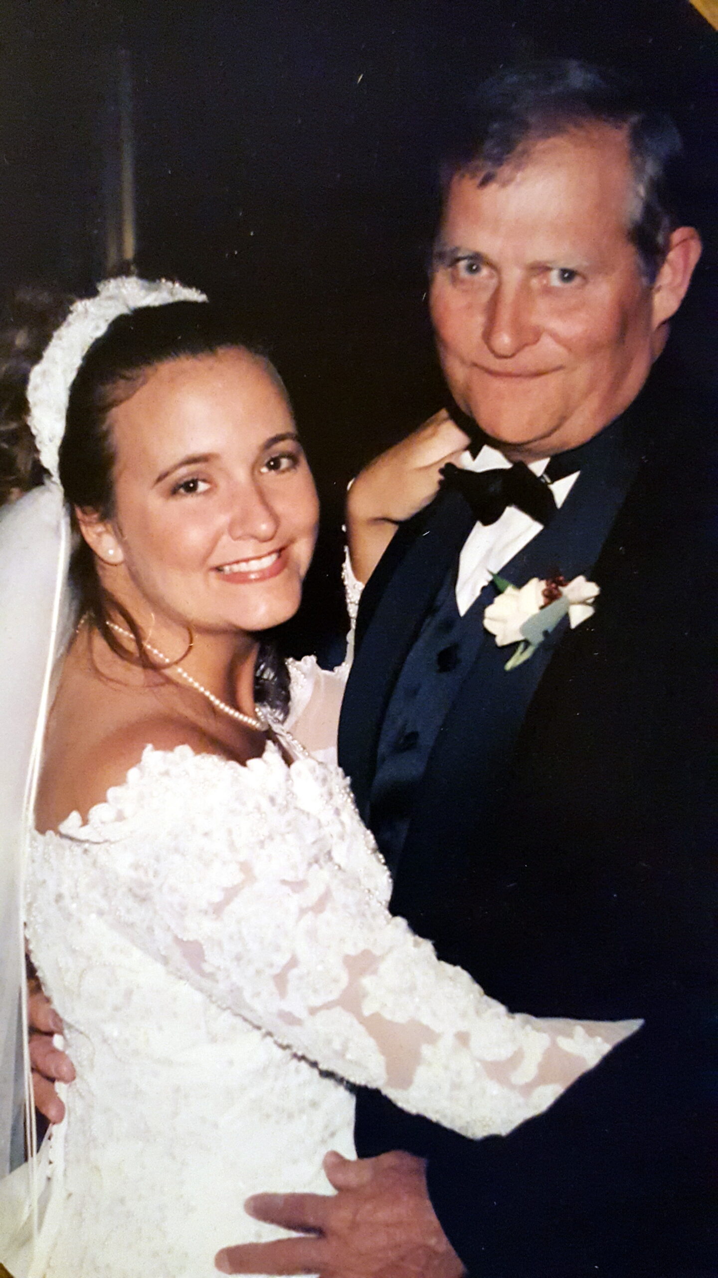 Bride in a white lace wedding dress smiles next to a man in a black tuxedo at a wedding.
