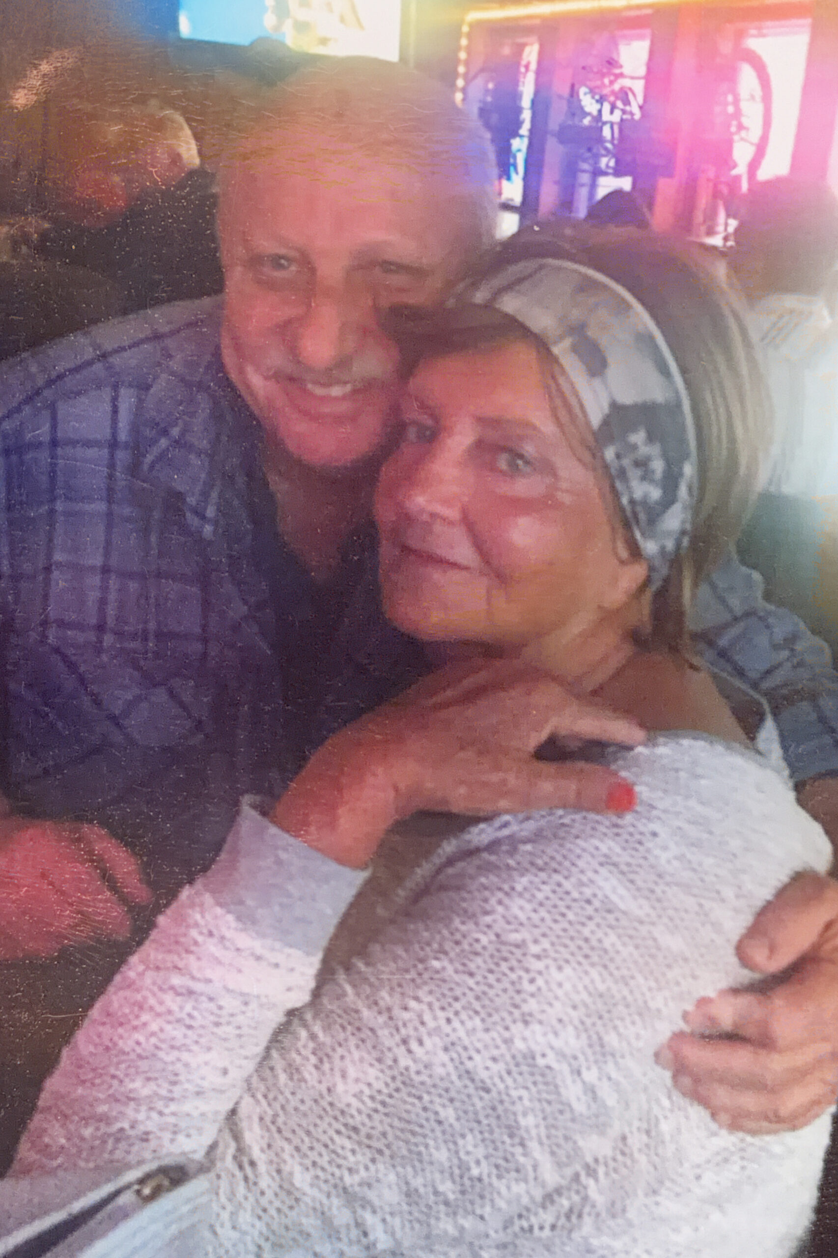 Two smiling adults share a tight hug in a warm indoor setting, looking at the camera.