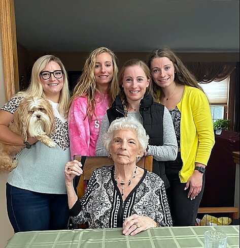 Grandmother seated at a table with four smiling young women and a small fluffy dog in a cozy home setting.