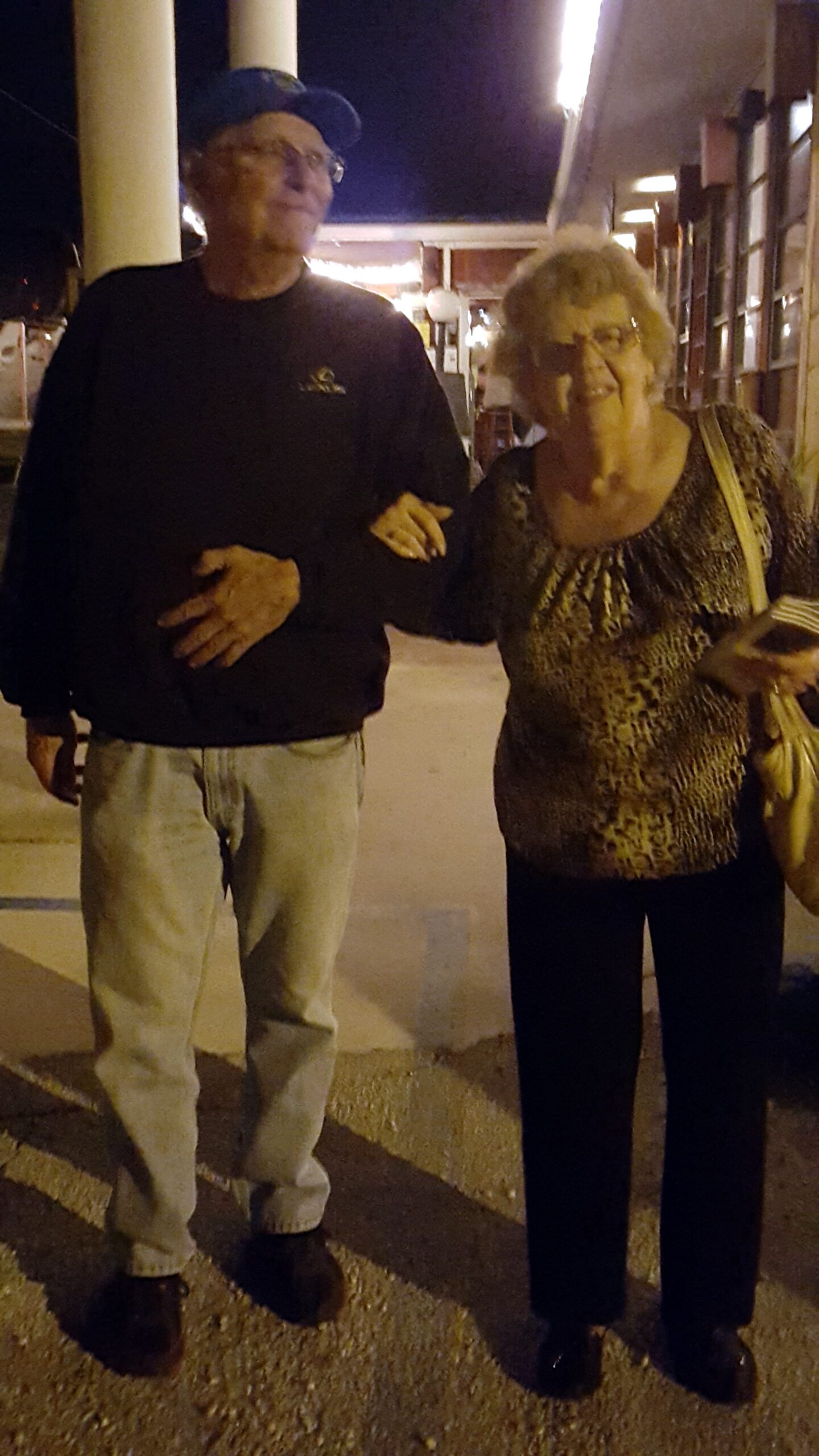 An elderly couple walking arm in arm on a nighttime street, smiling at the camera.