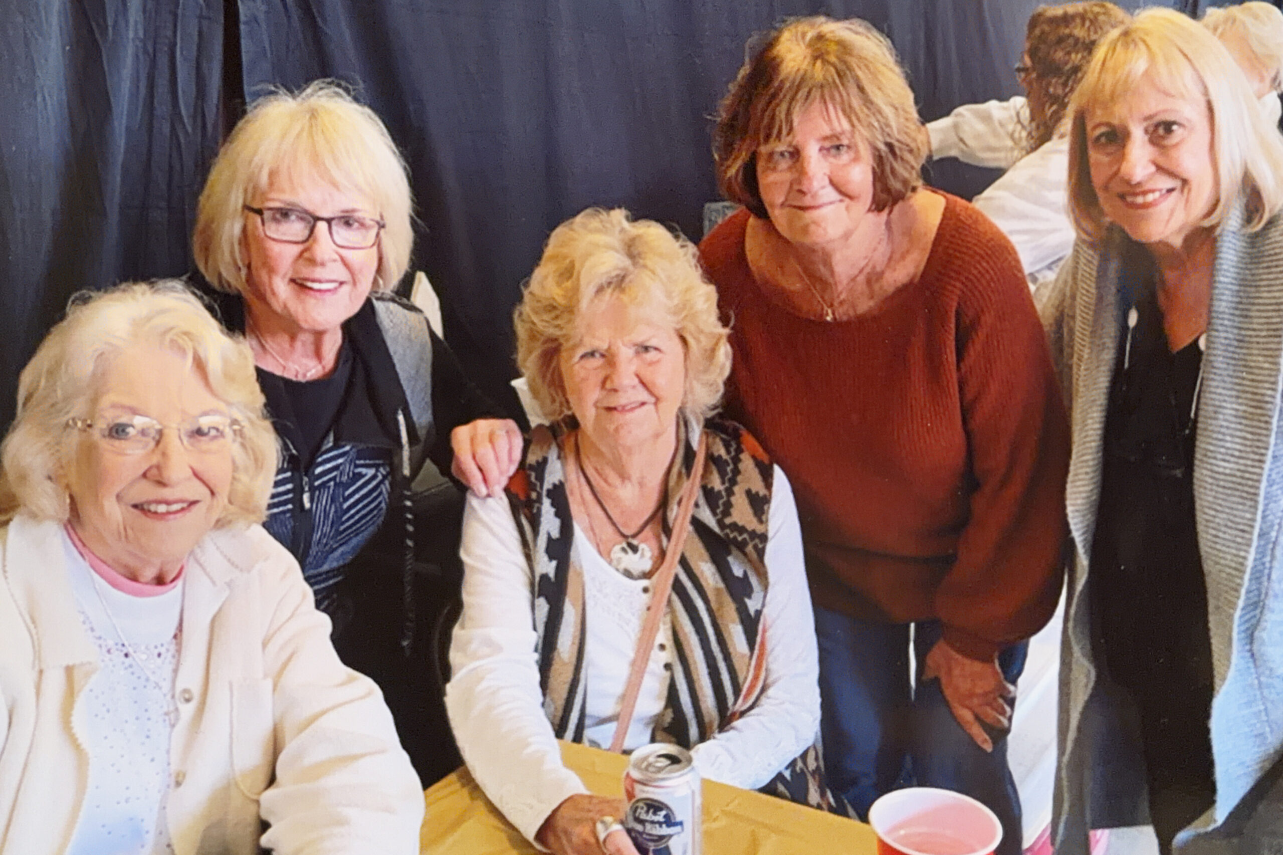 Group of smiling older women gathered around a table with drinks at a casual get-together.