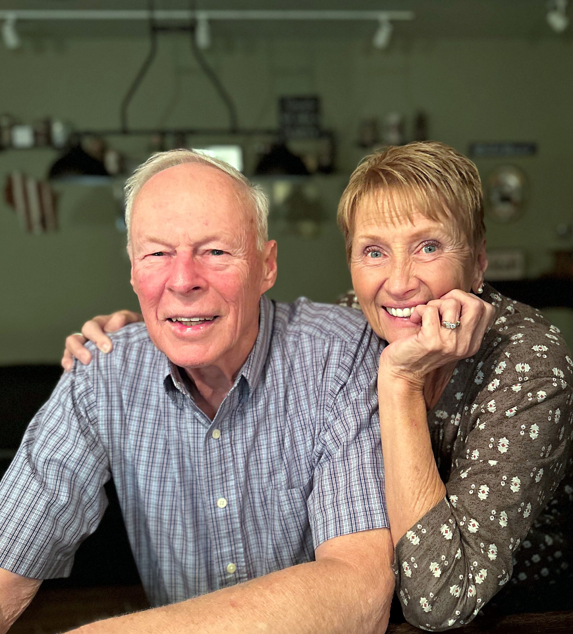Two elderly adults smile at the camera, arms around each other in a warm embrace inside a home setting.