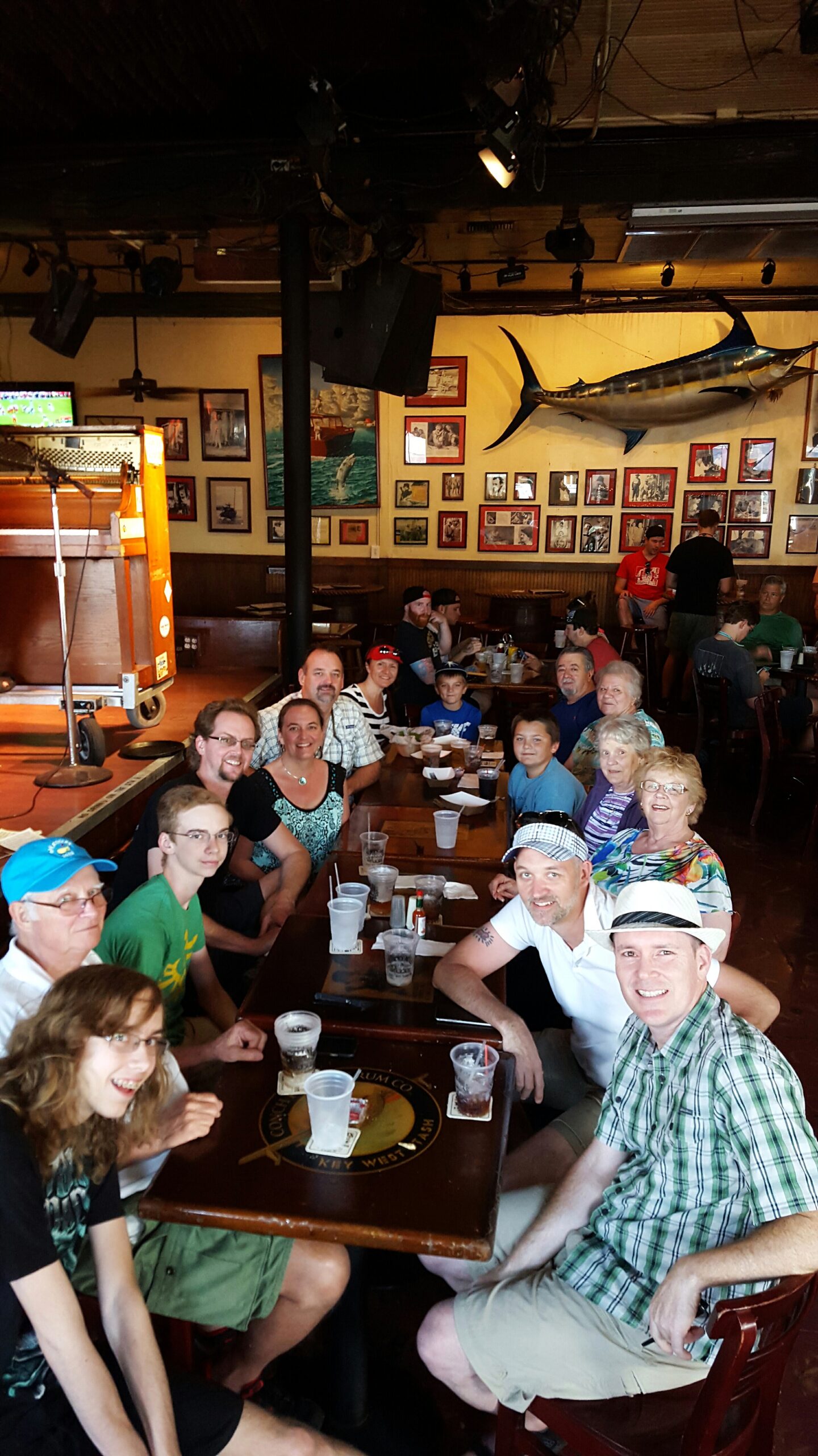 Large group of people sitting around a long restaurant table, smiling and enjoying drinks, with walls full of framed photos and a mounted marlin overhead.