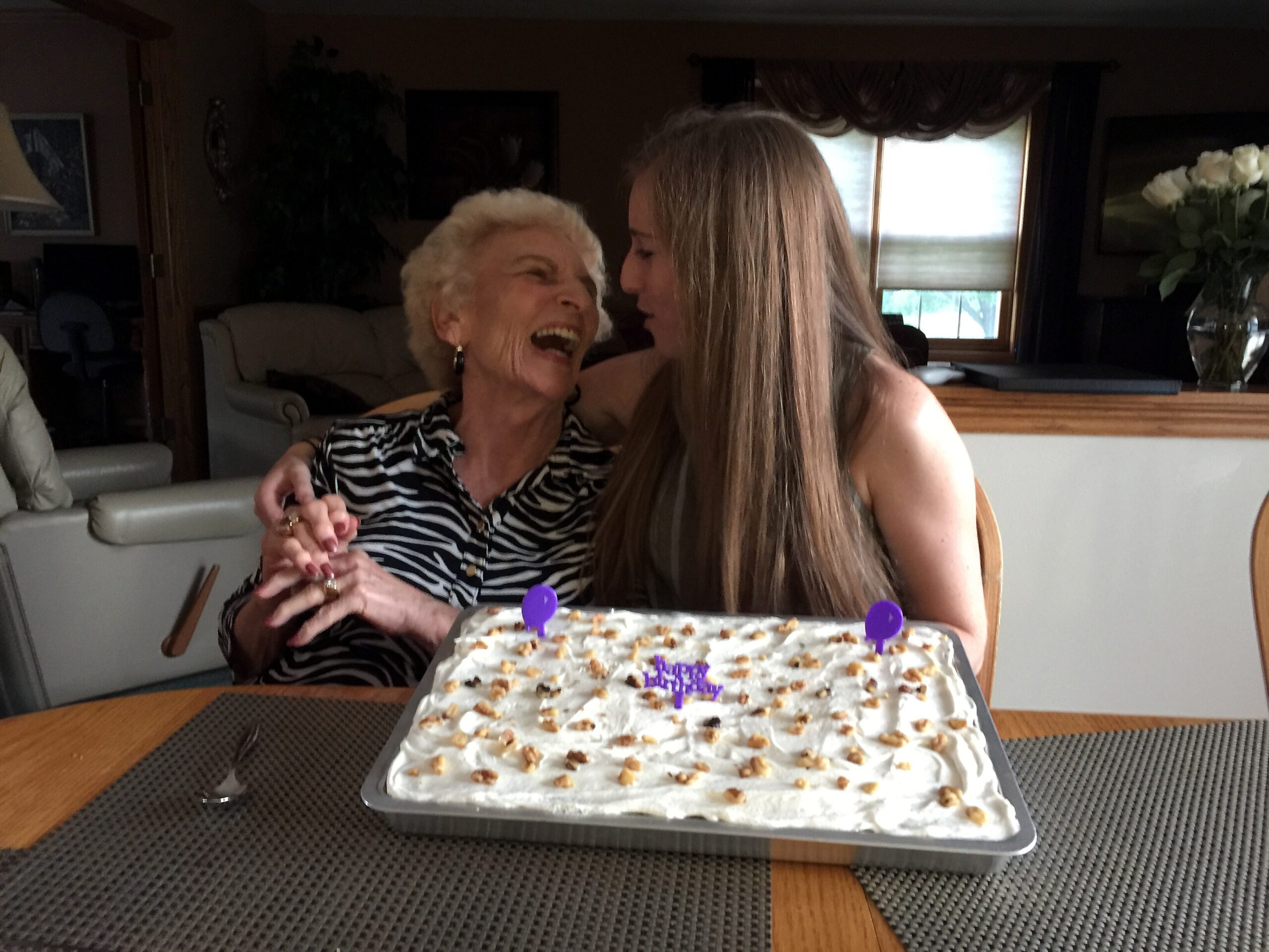 Two women share a joyful moment at a birthday with a large frosted cake in front of them.