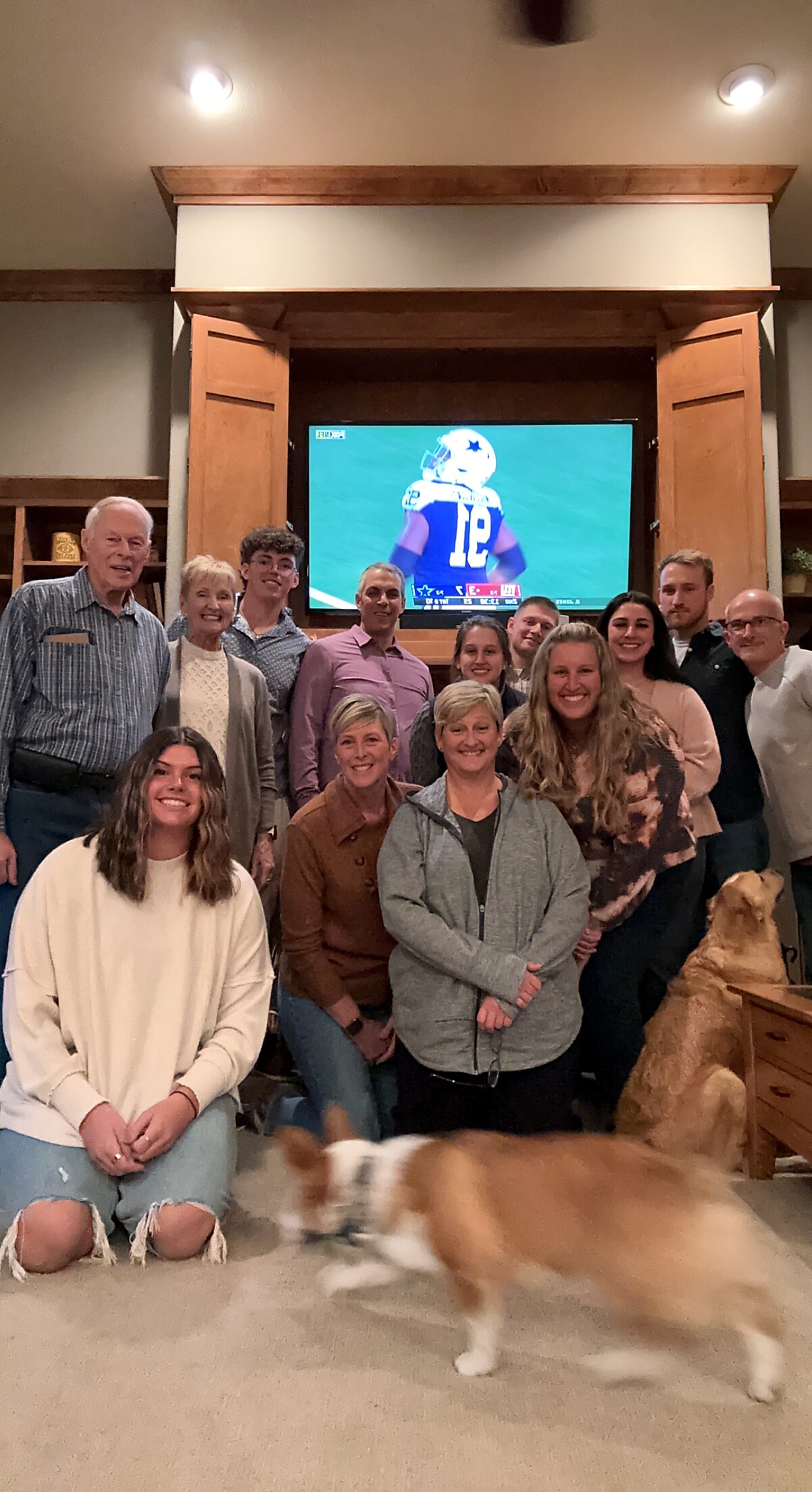 Group of people posing indoors in a living room with a football game on the TV screen behind them; a dog is moving in the foreground.