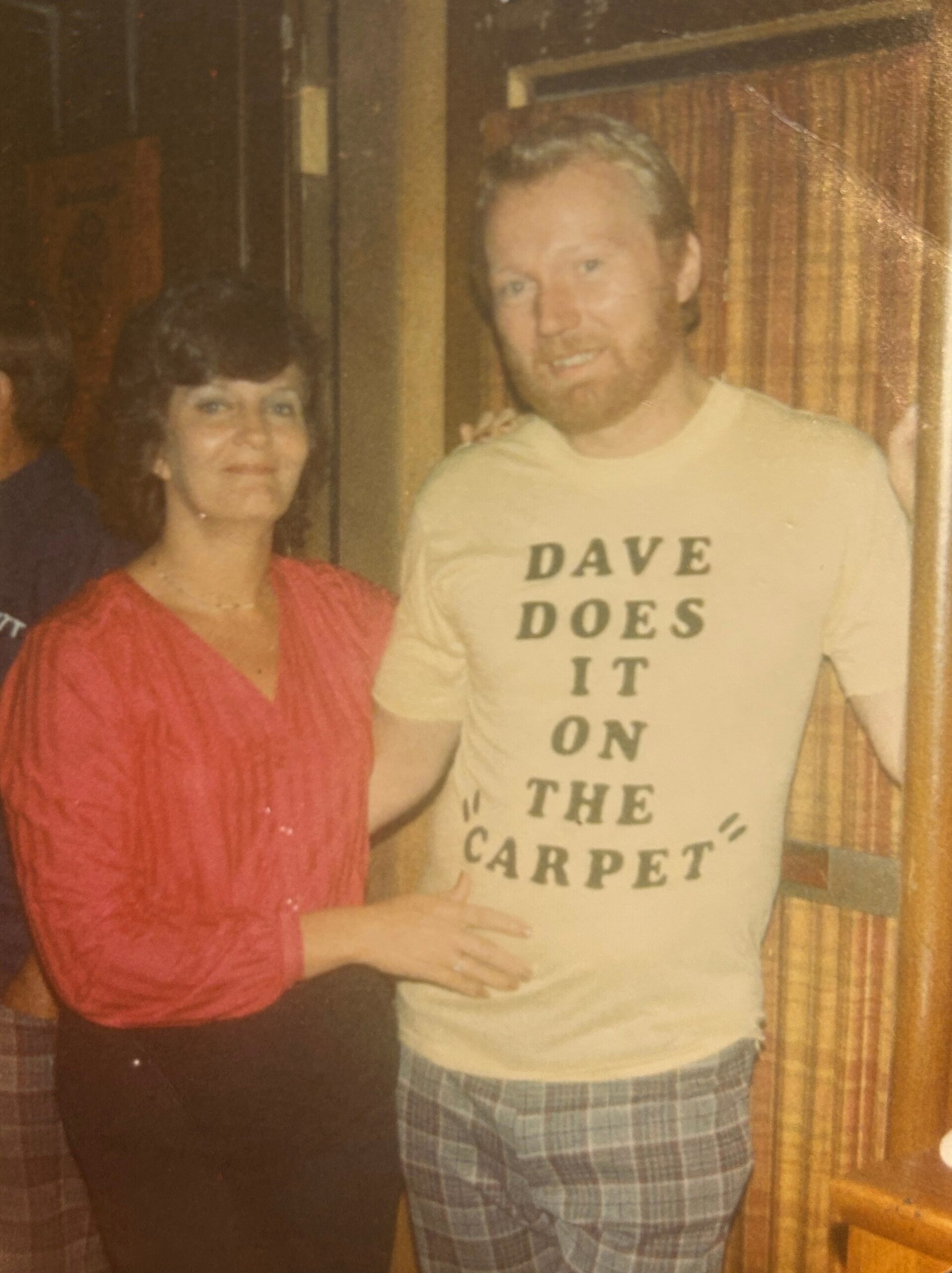 Smiling woman in a red blouse stands beside a man in a light T‑shirt with bold text, indoors by wood-paneled walls, both posing for a photo.
