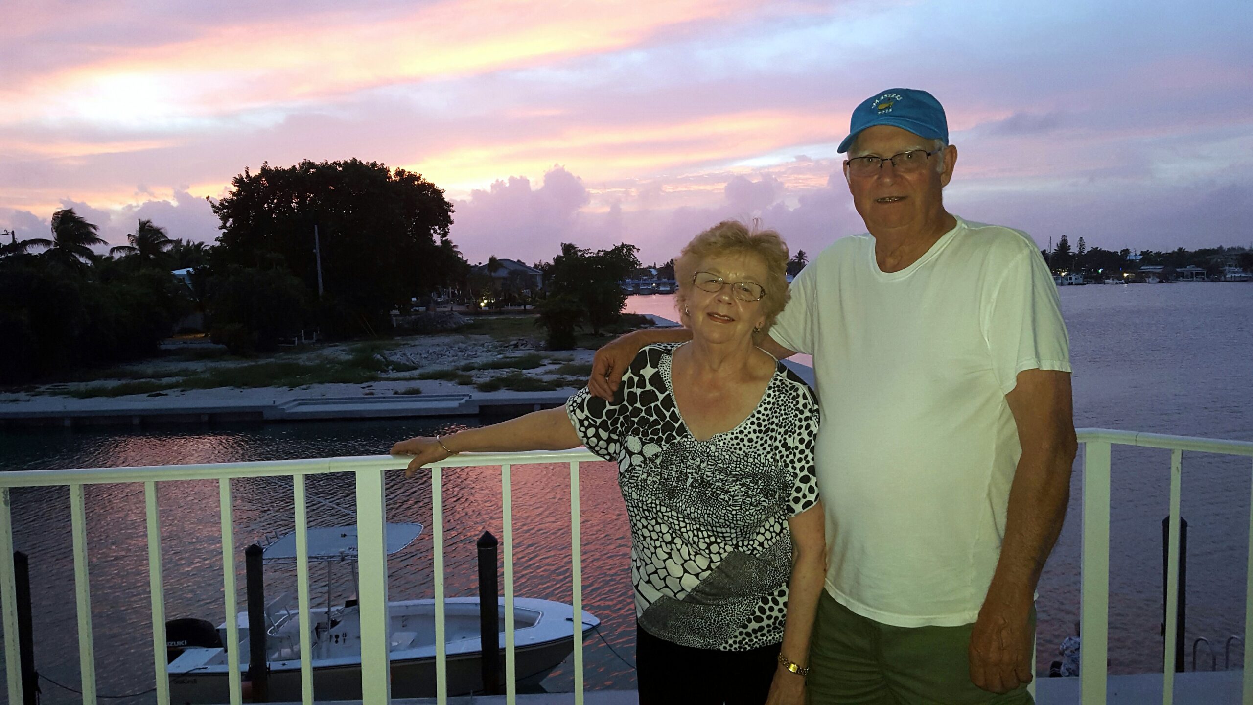 Older couple leaning on a railing by a marina at sunset, boats on the water and pink-orange sky behind them.