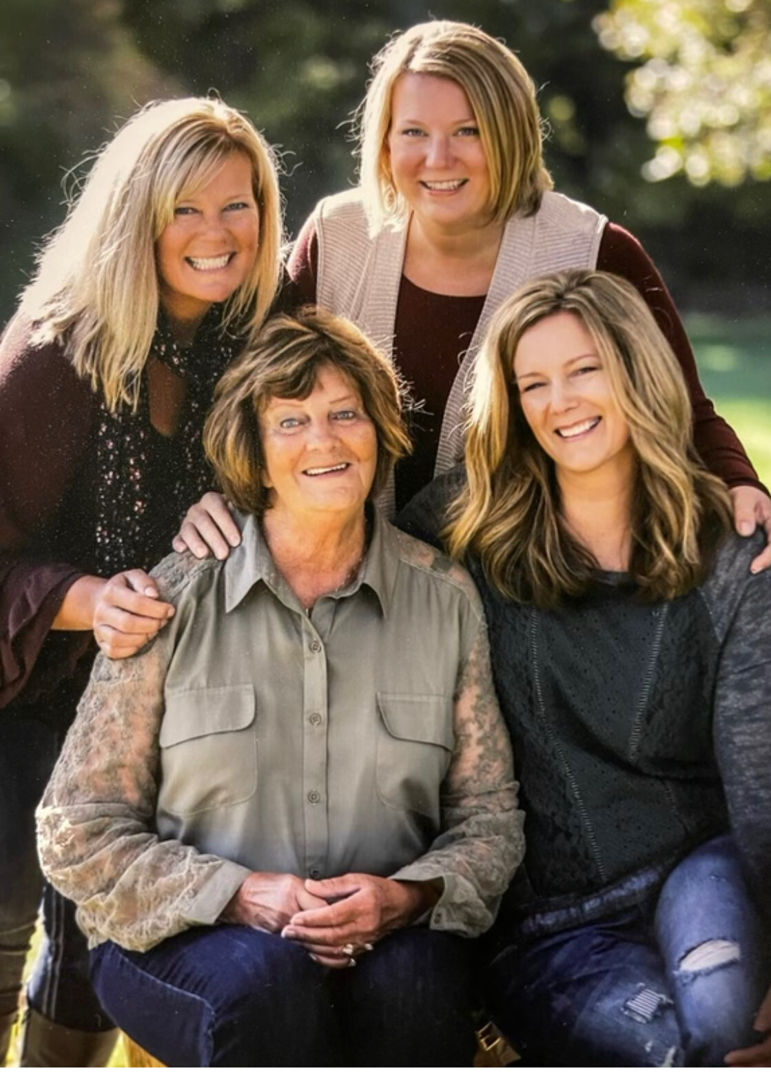 Four women pose for a cheerful outdoor group photo, with one seated in front center and three standing behind her.