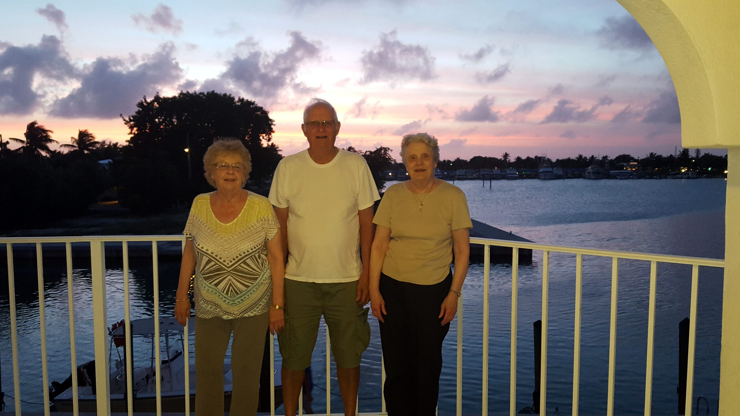 Three seniors stand side by side on a railing overlooking a marina at sunset, with boats in the distance.