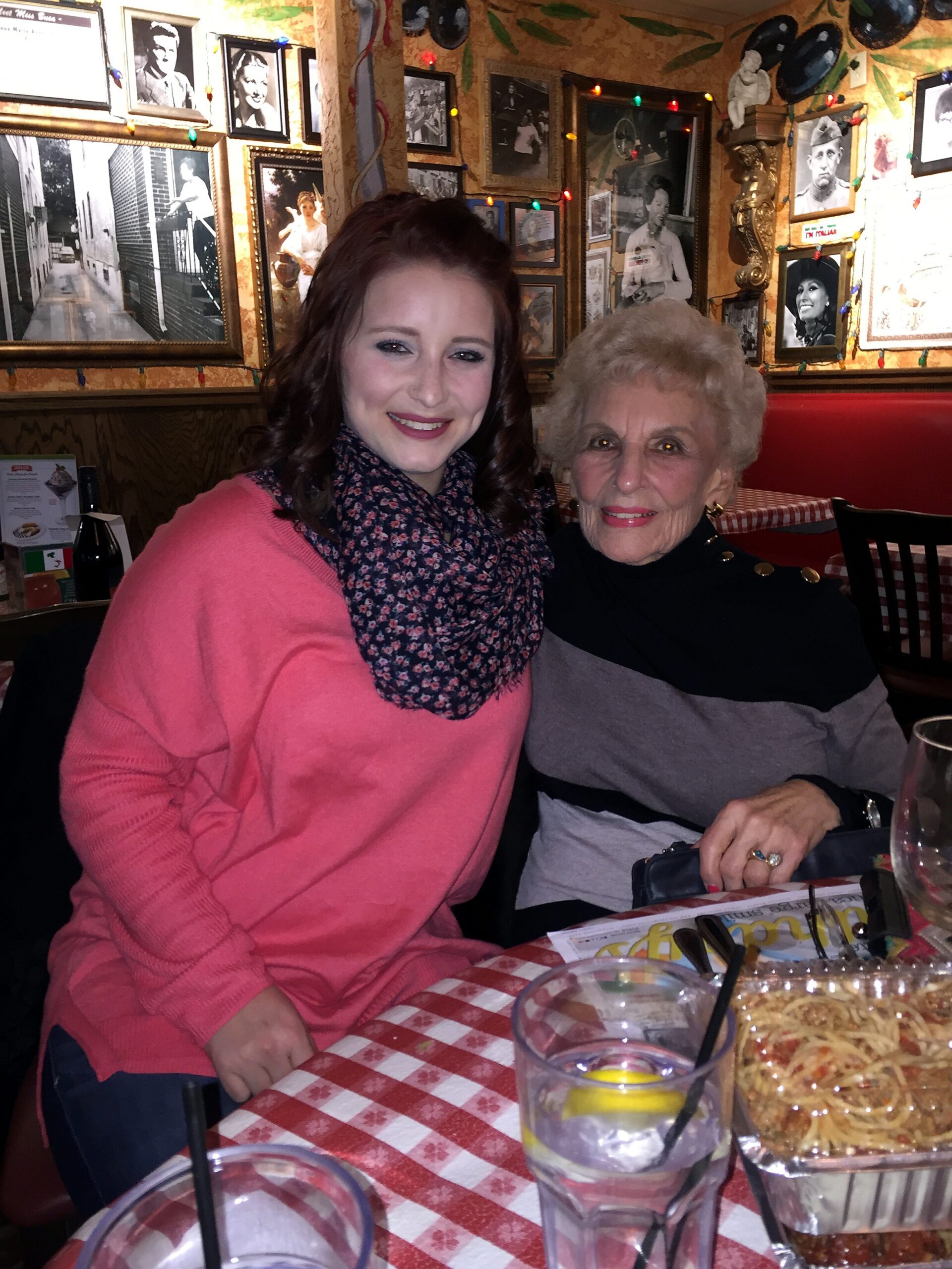 Two women smiling at a table in a cozy restaurant filled with black-and-white photos on the wall; one wearing a pink sweater and floral scarf.