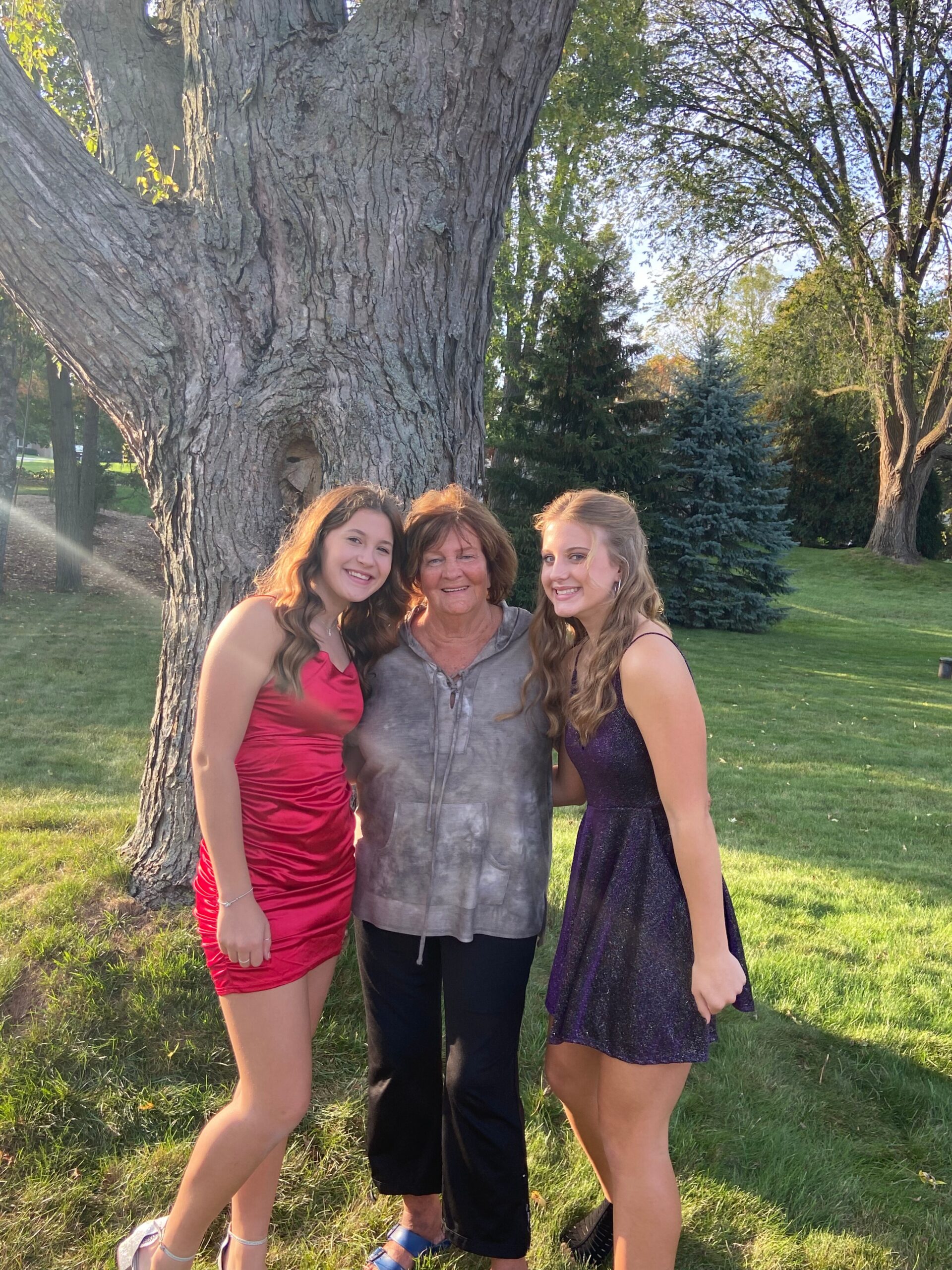Three women posing together by a large tree in a sunny park: an older woman in the center flanked by two younger women in red and purple dresses, all smiling.