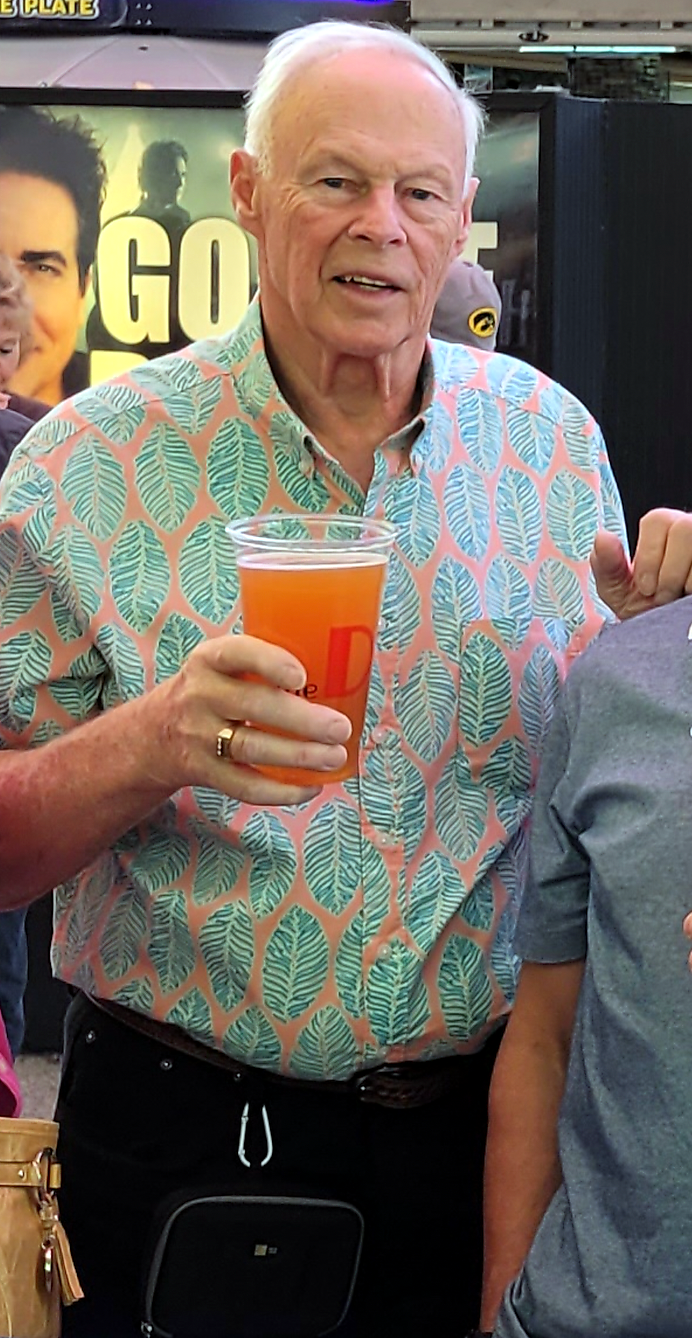 Older man with white hair in a pink-and-teal leaf-pattern shirt, holding an orange drink in a clear plastic cup.