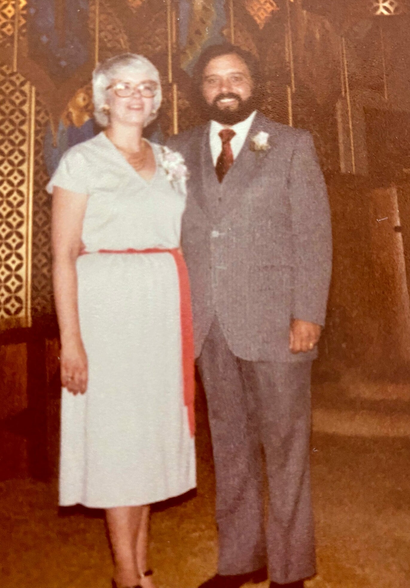Smiling couple posing together at a formal event: woman in a light dress with a red sash and boutonniere, man in a gray suit with a tie and boutonniere.