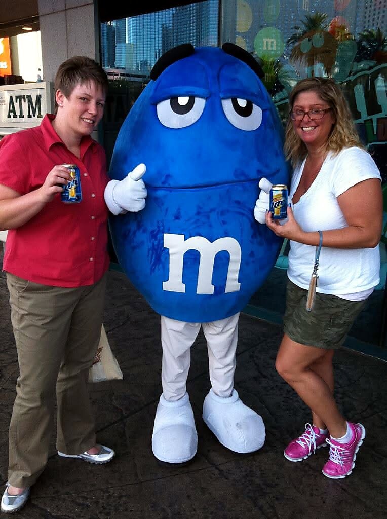 Two people pose with a large blue M&M mascot outside a storefront, each holding a can of soda.