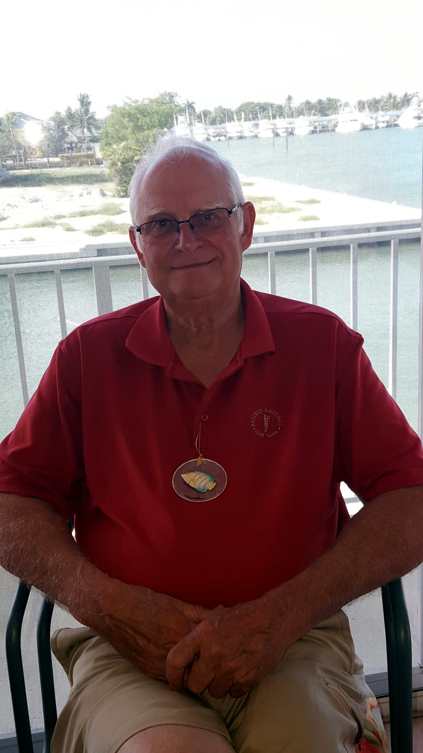 Older man with white hair and glasses, wearing a red polo, sitting on a balcony by the water with boats in the background.