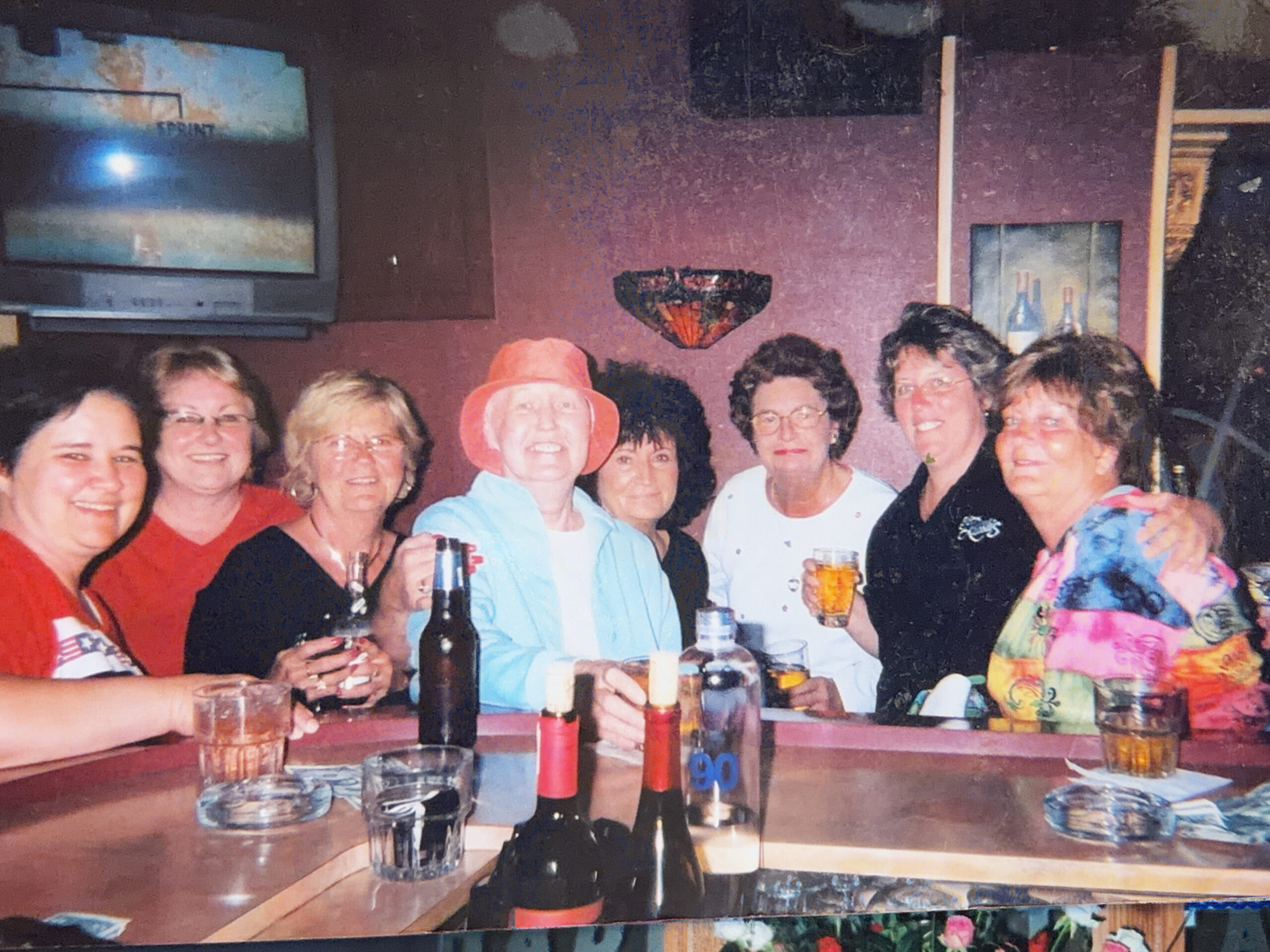 Group of adults gathered around a bar table, smiling at the camera with drinks and bottles on the table in front of them.
