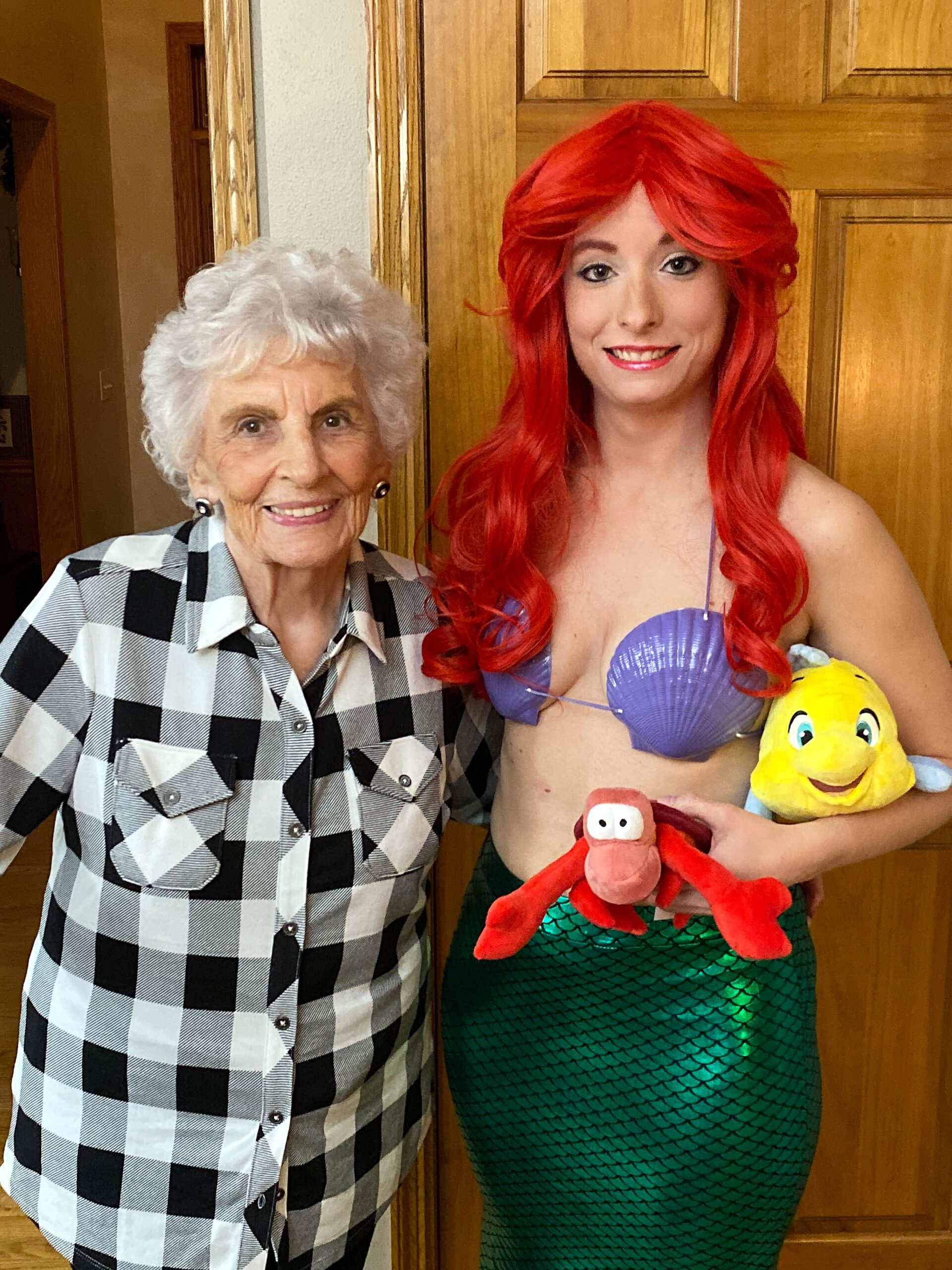 Smiling elderly woman in a black-and-white checkered shirt standing beside a person in a red-mermaid costume holding stuffed sea creatures (crab and duck).