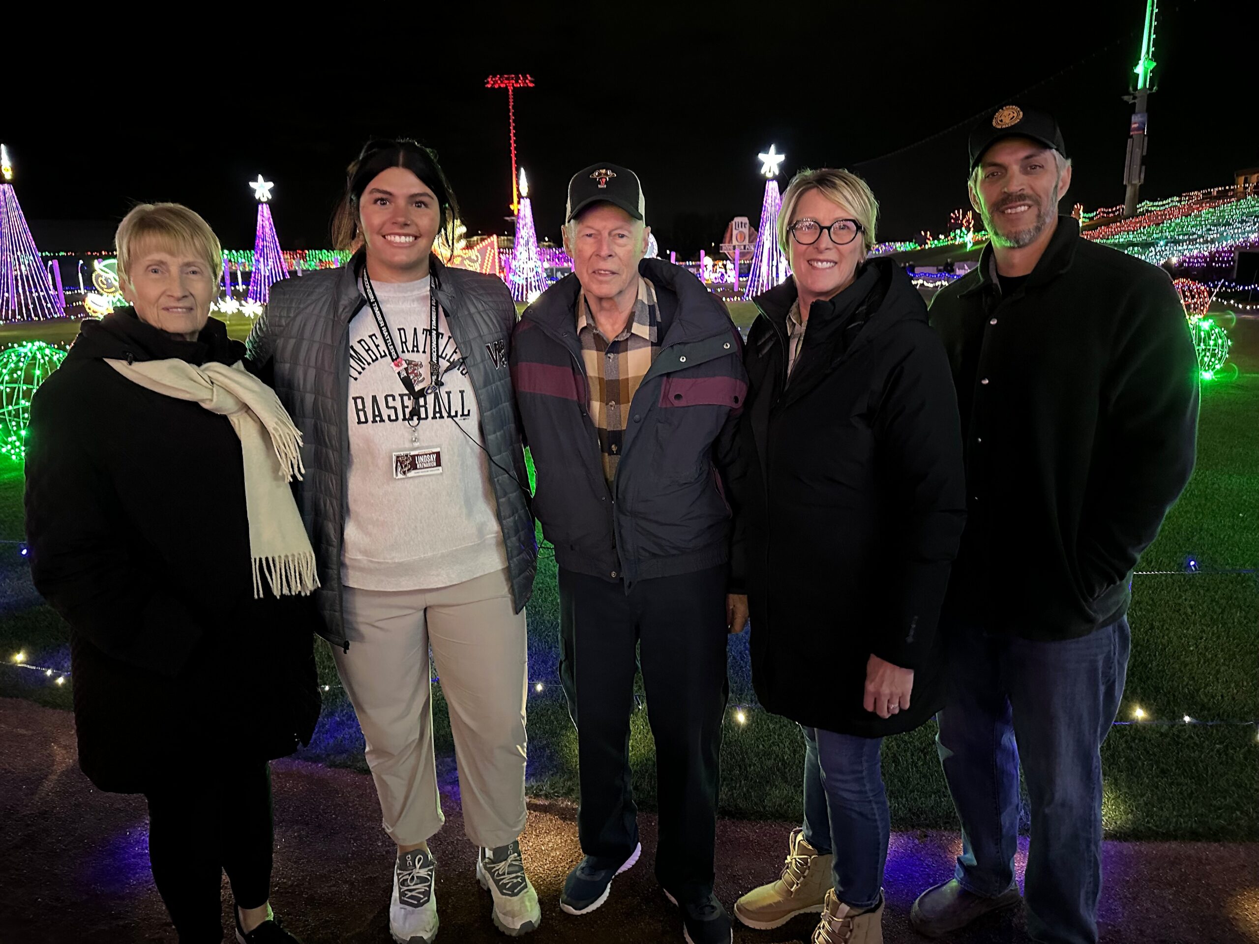 Five adults posing together at a nighttime holiday light display, smiling at the camera in front of colorful festive lights in a park.