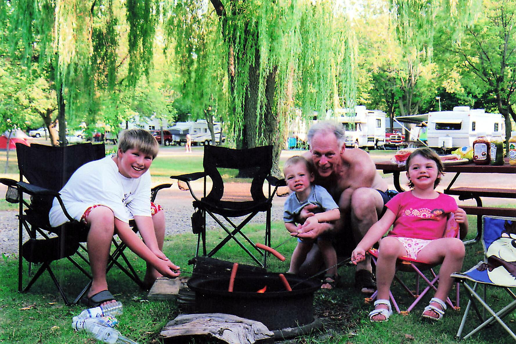 Family at a campground enjoying an outdoor campfire, roasting marshmallows in a grassy park with trees and RVs in the background.