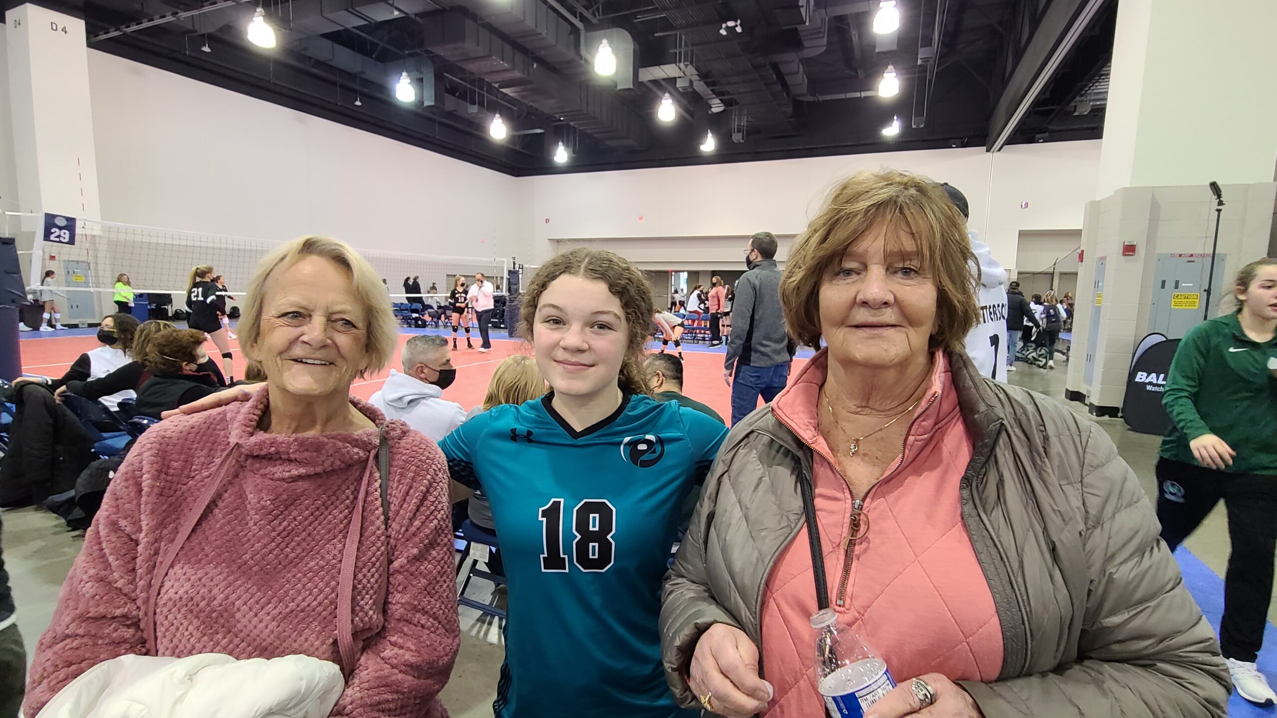 Three women/teen posing for a photo at an indoor volleyball event, with players and a net visible in the background.