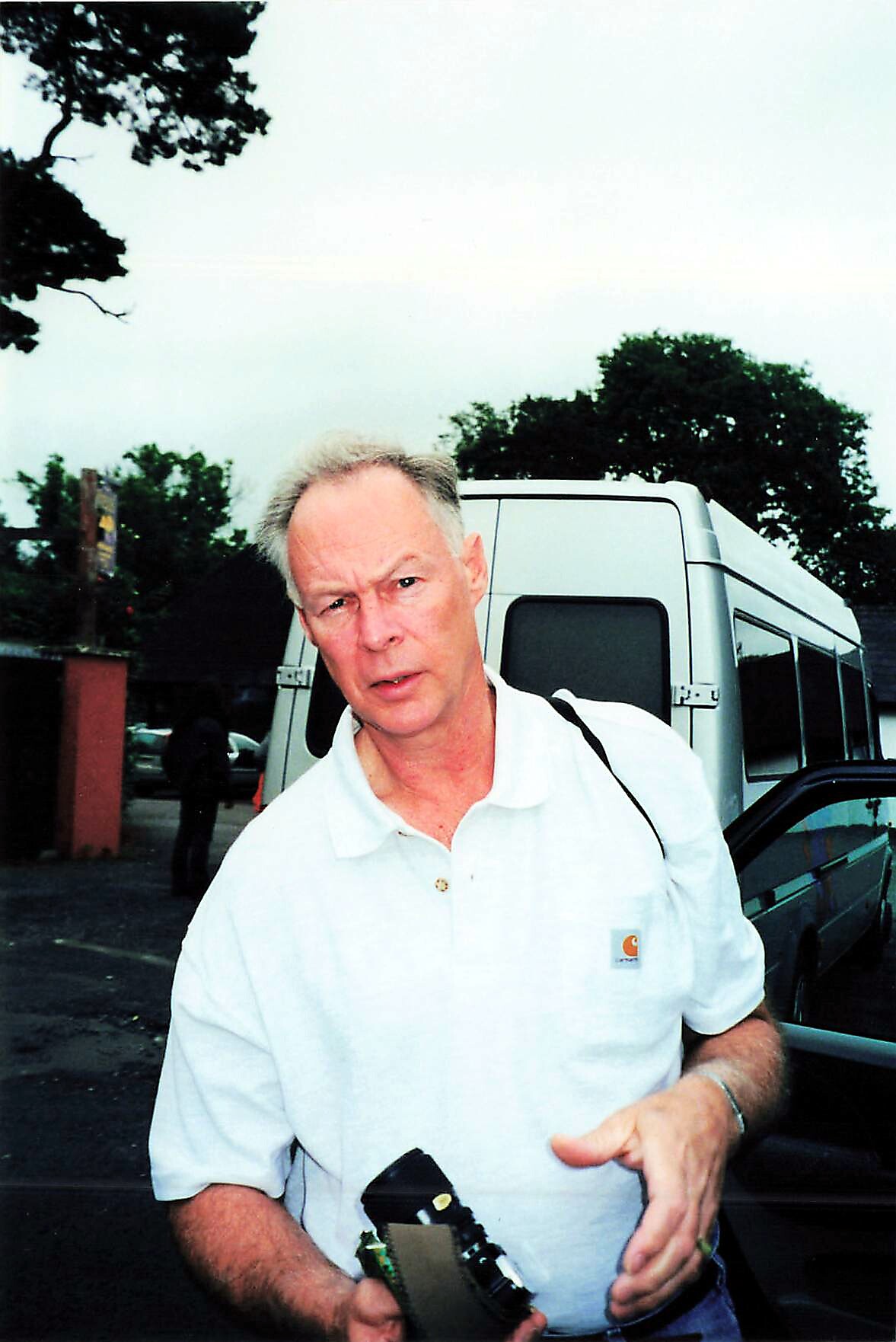Older man in a white polo shirt stands beside a silver van in a parking lot, holding a small camera or device in his hands beside a black bag/strap.