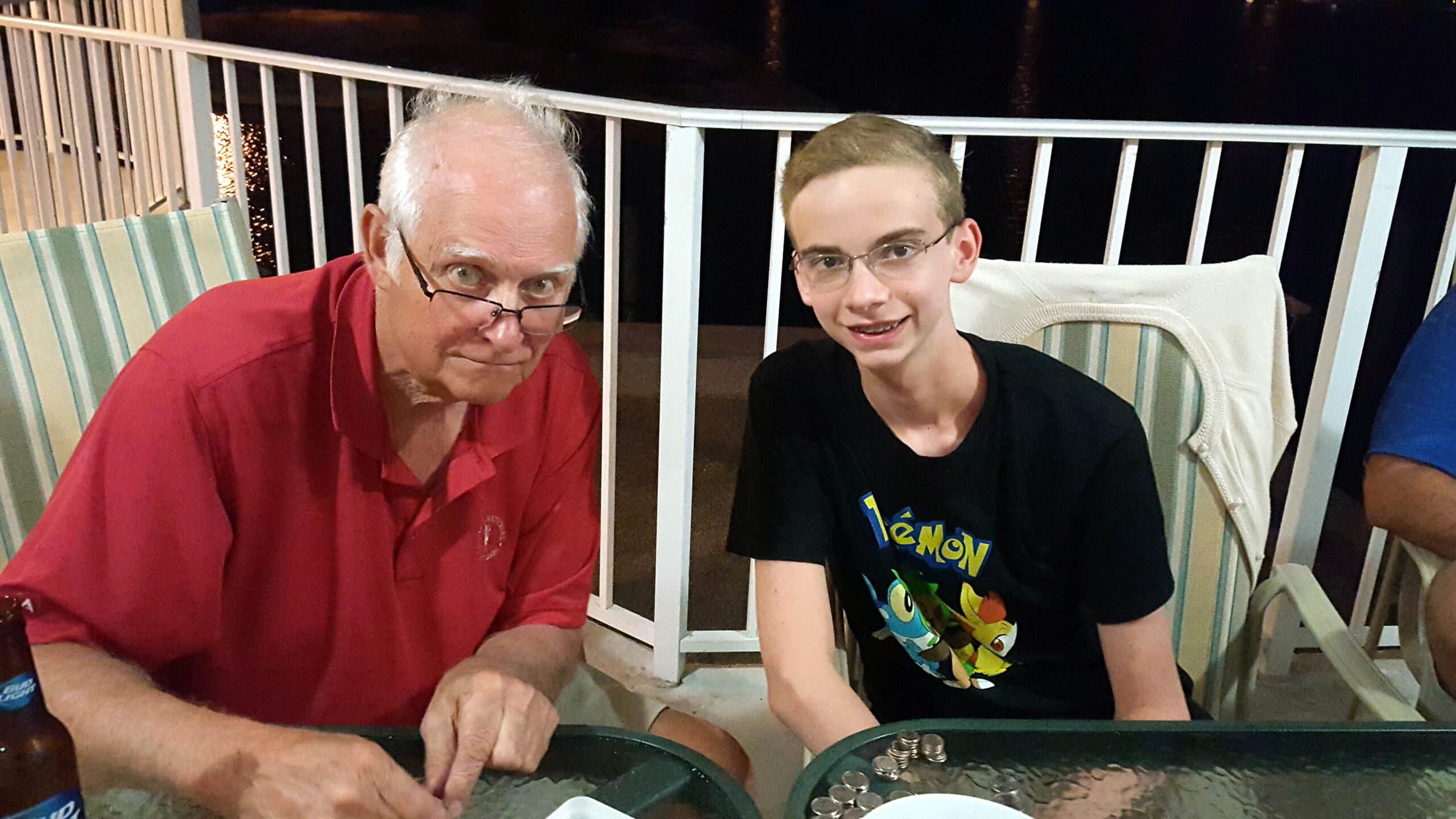 Two men sit at a table on a balcony at night, sorting coins in trays; an older man in a red polo and a younger man with glasses in a Pokémon shirt.