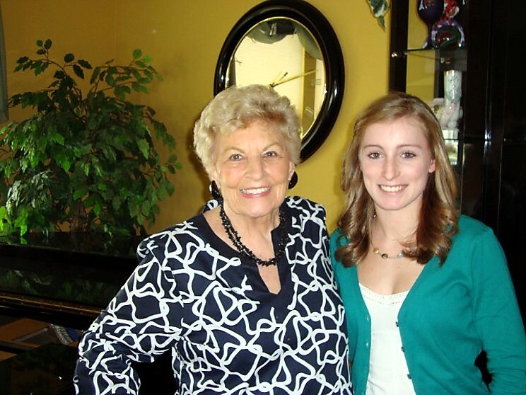 Two smiling women pose indoors: an older woman in a black-and-white patterned blouse and a younger woman in a teal cardigan, standing side by side in front of a plant and round mirror.