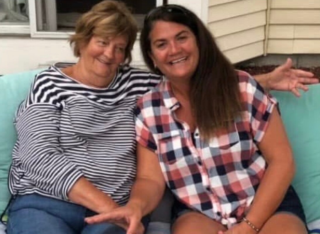Two women sit close together on a light-blue couch, smiling at the camera outside a house.