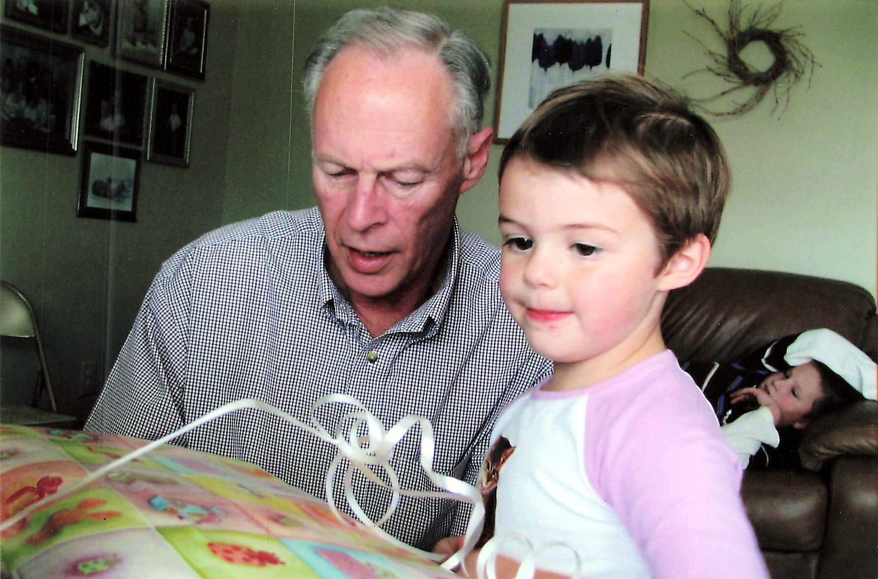 Older man and a young girl sit at a table opening a colorful gift together in a home living room, with a child on a sofa in the background.