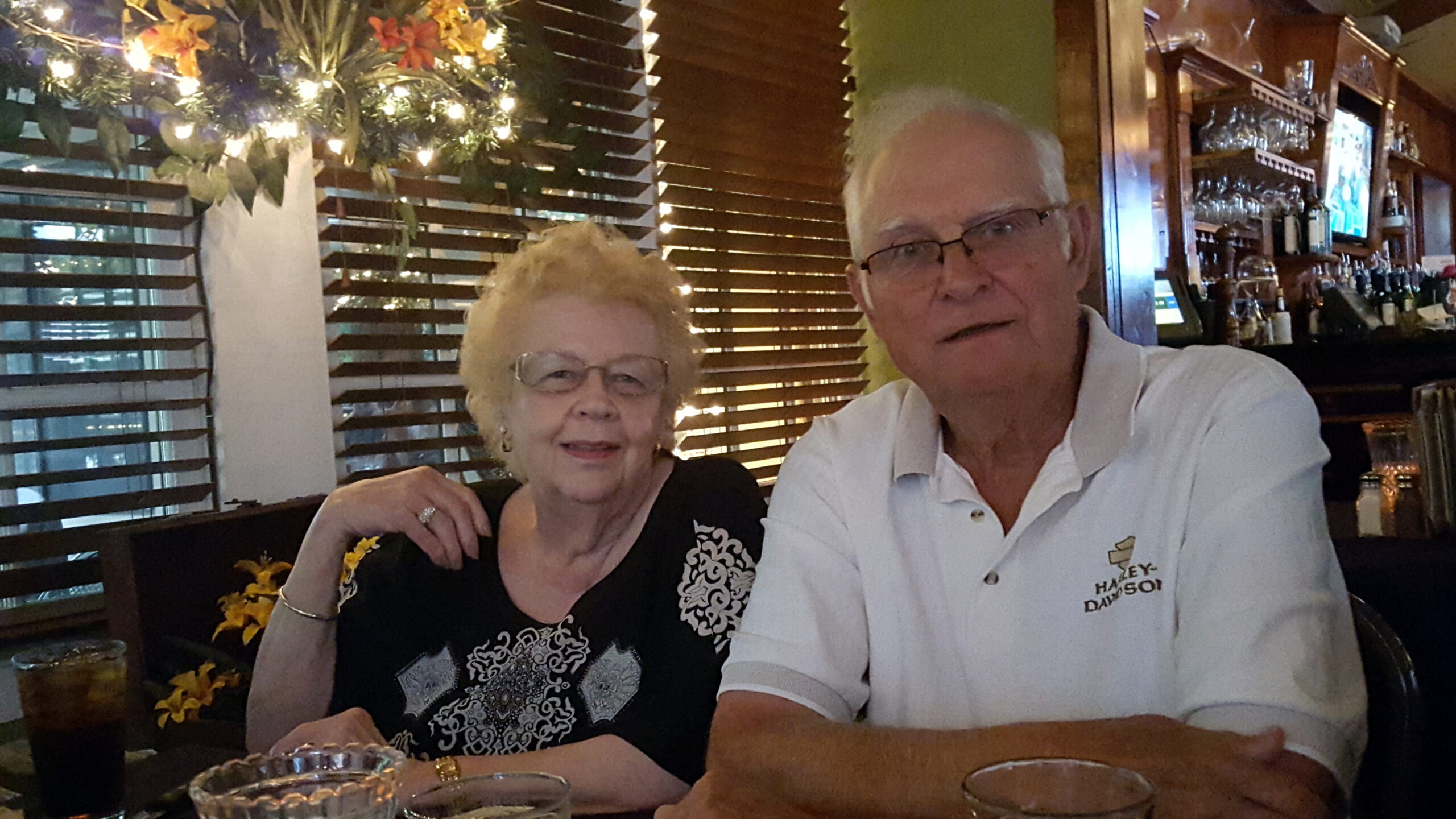 Older couple at a restaurant bar posing for the camera; drinks on the table, warm string lights behind them.