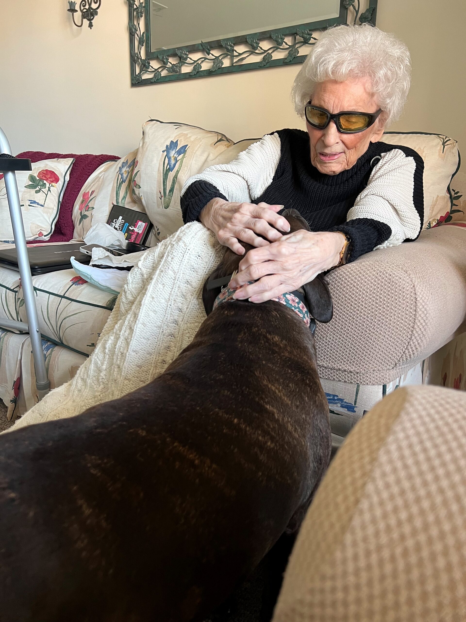 Older woman with white hair and yellow-tinted sunglasses sits on a floral sofa, smiling as a large brown dog sits close and rests its head near her lap with a blanket draped over them.