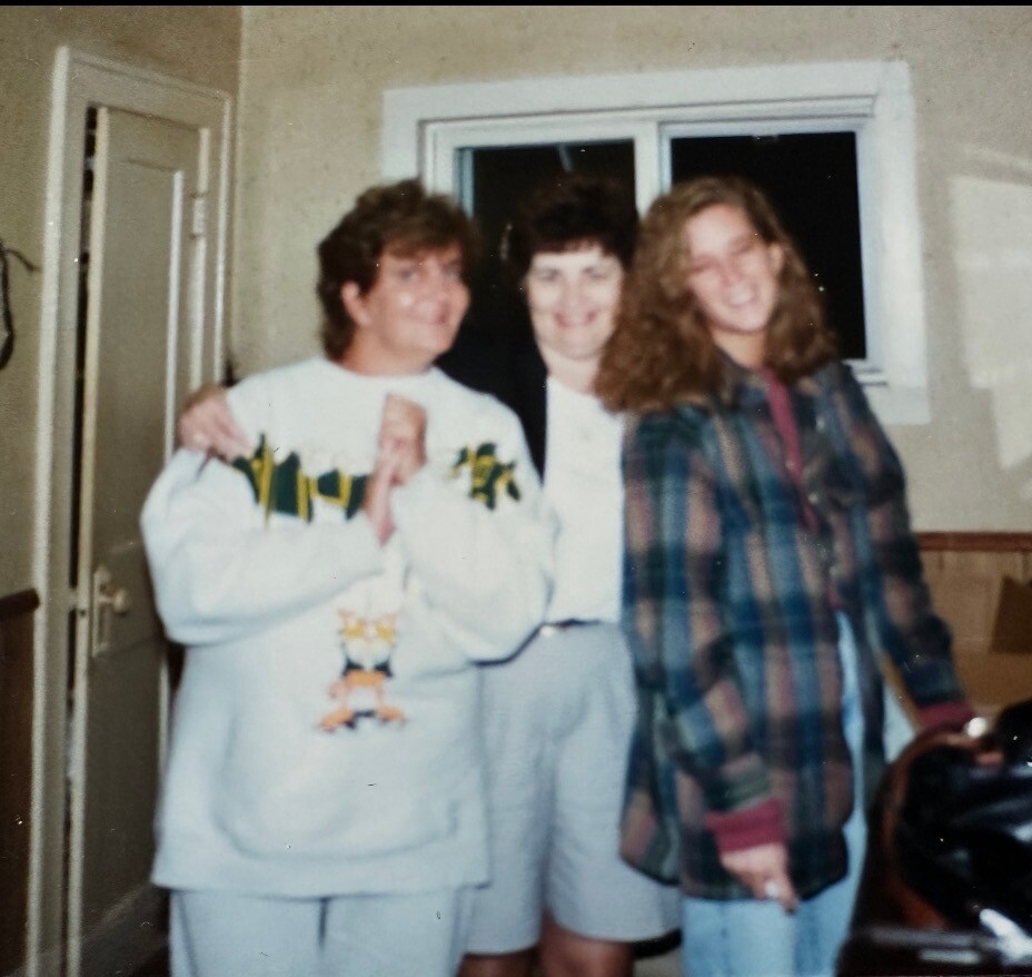 Three friends posing for a casual photo indoors, standing close together near a doorway; left person in a light sweatshirt with a graphic, middle person in a white top, right person in a plaid jacket.
