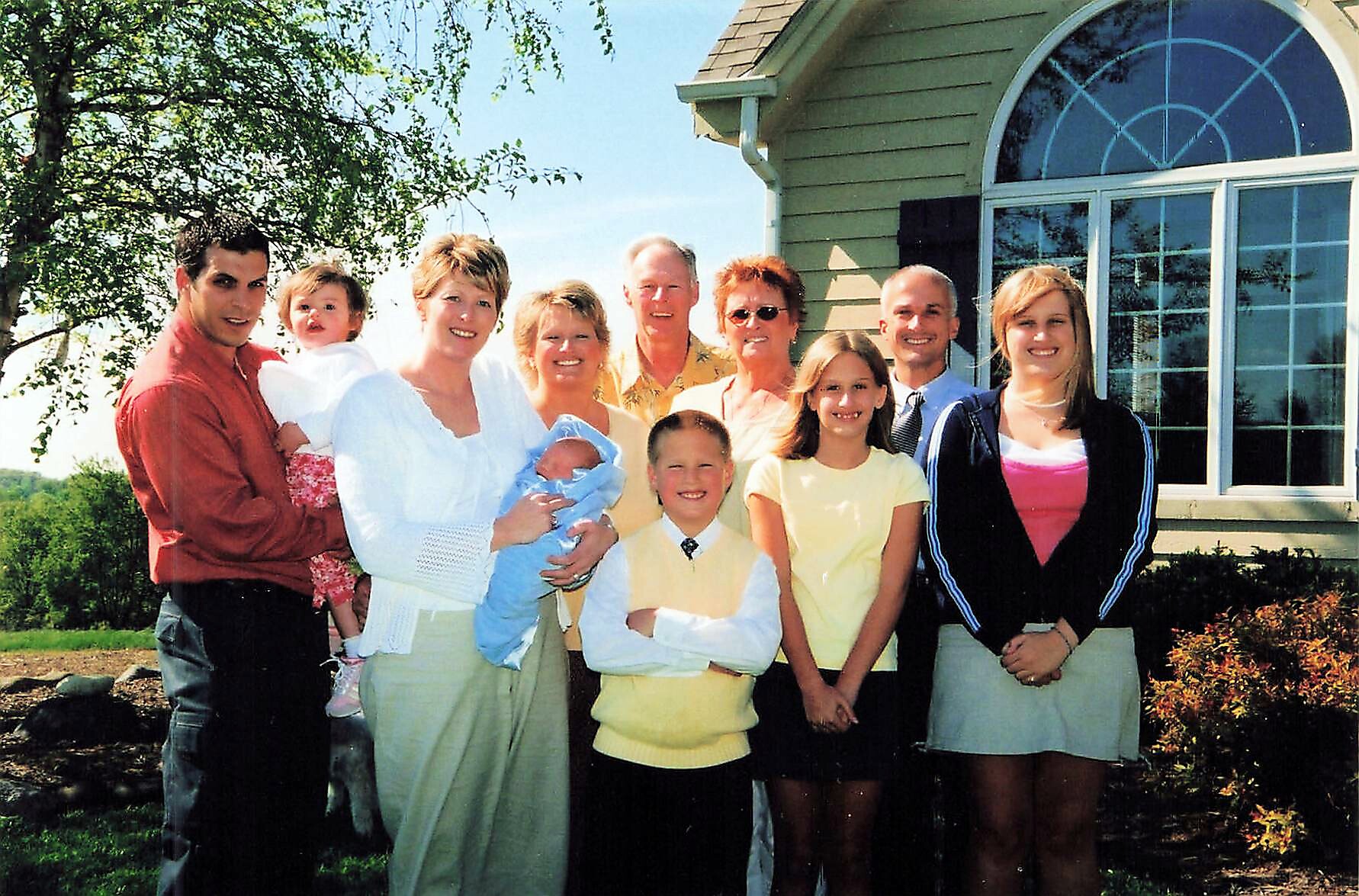 Multi‑generational family poses for a portrait on the yard in front of a green house, with adults and several children, including a baby wrapped in blue, all smiling.