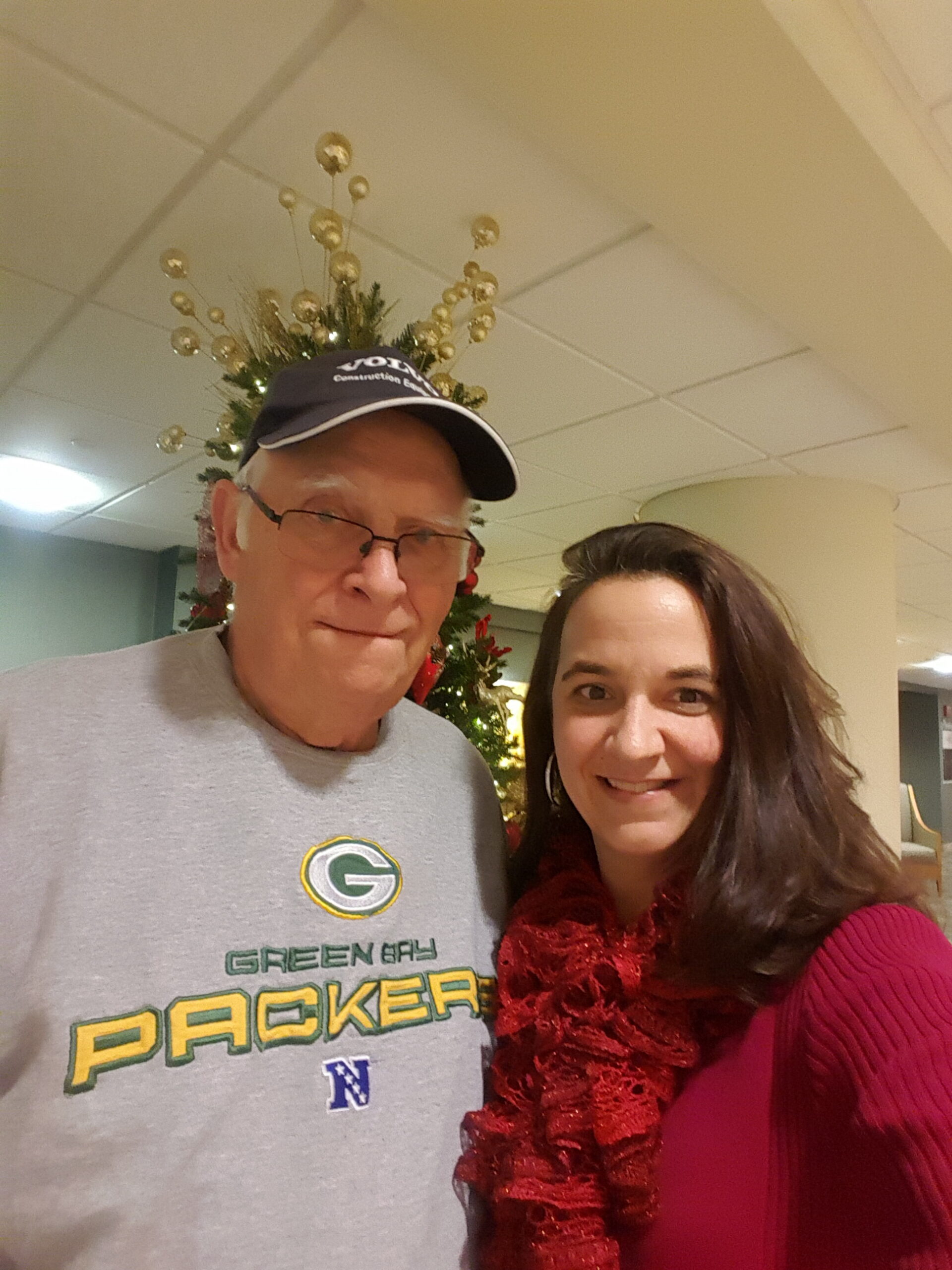 Two people take a selfie indoors in front of a Christmas tree; man wears a Green Bay Packers shirt and cap, woman in pink with a red scarf.